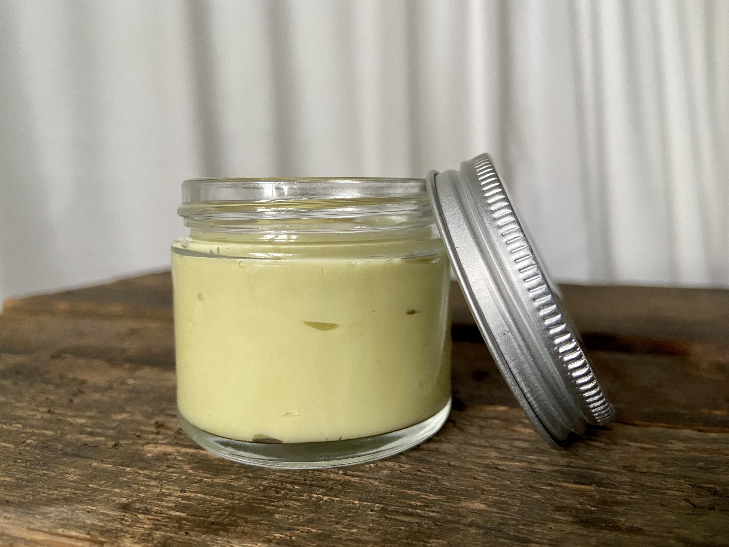 Image of glass jar containing sage green salve on wooden surface with white fabric in background.