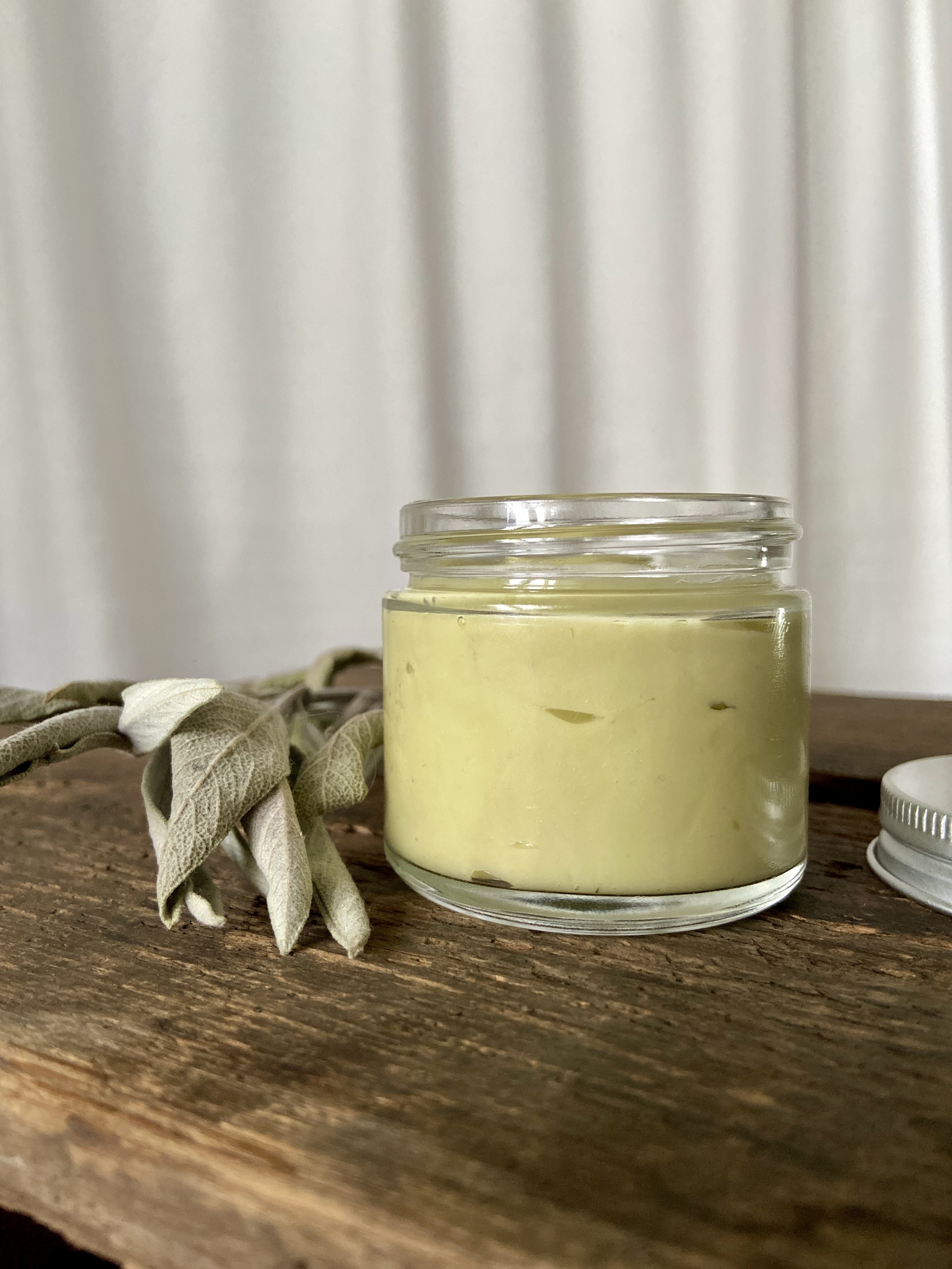 Image of glass jar containing sage green salve on wooden surface with white fabric in background. There is a spring of sage next to the glass jar