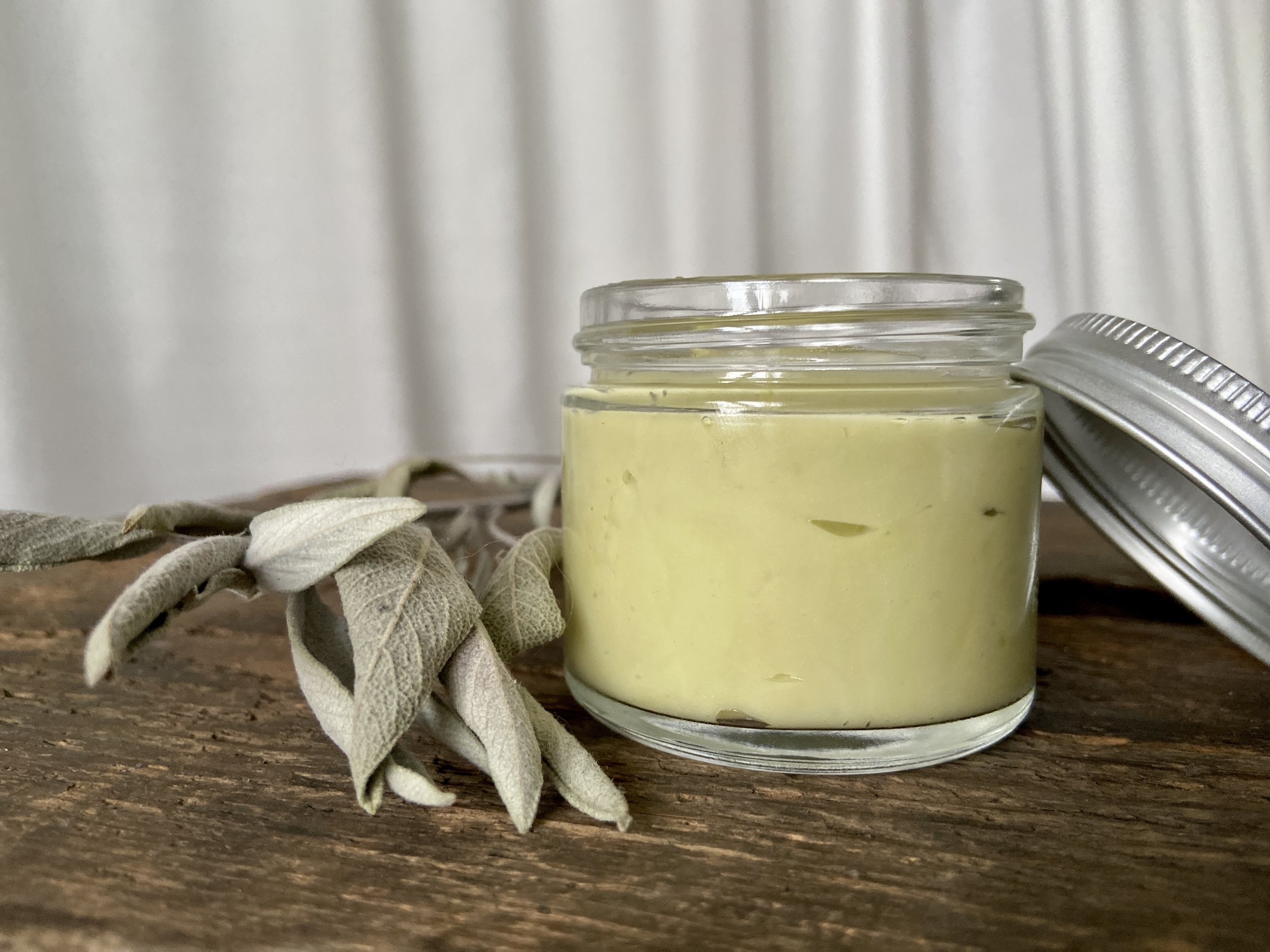 Image of glass jar containing sage green salve on wooden surface with white fabric in background. There is a spring of sage next to the glass jar