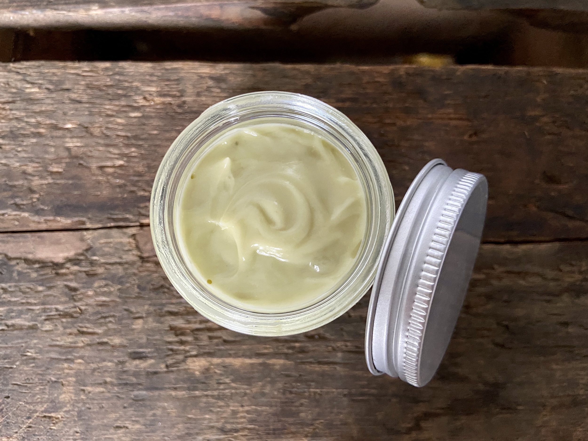 Image of glass jar containing sage green salve on wooden surface with white fabric in background.