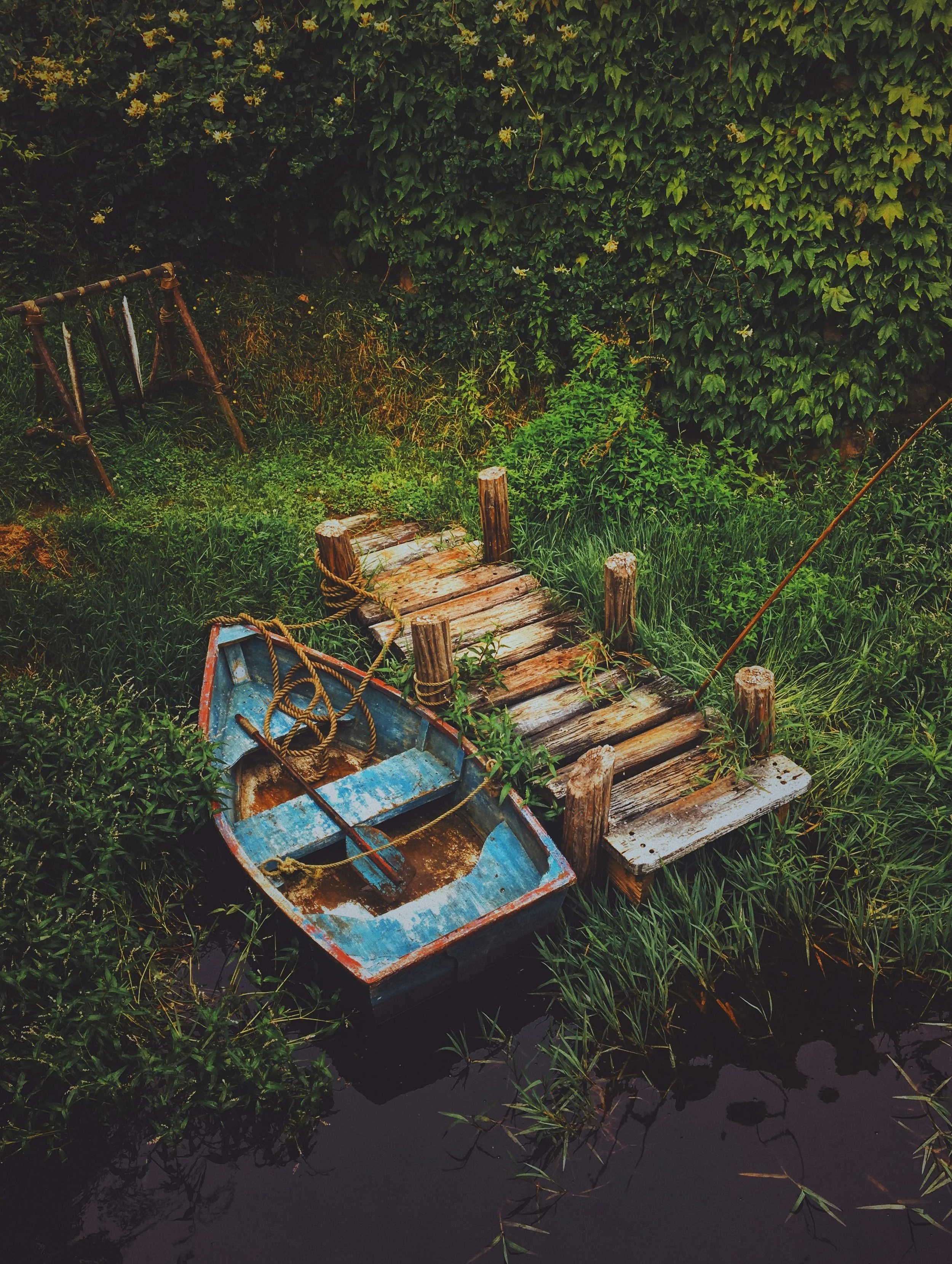 A miniature wooden boat is tied to a miniature wooden dock. They are both resting on grass.