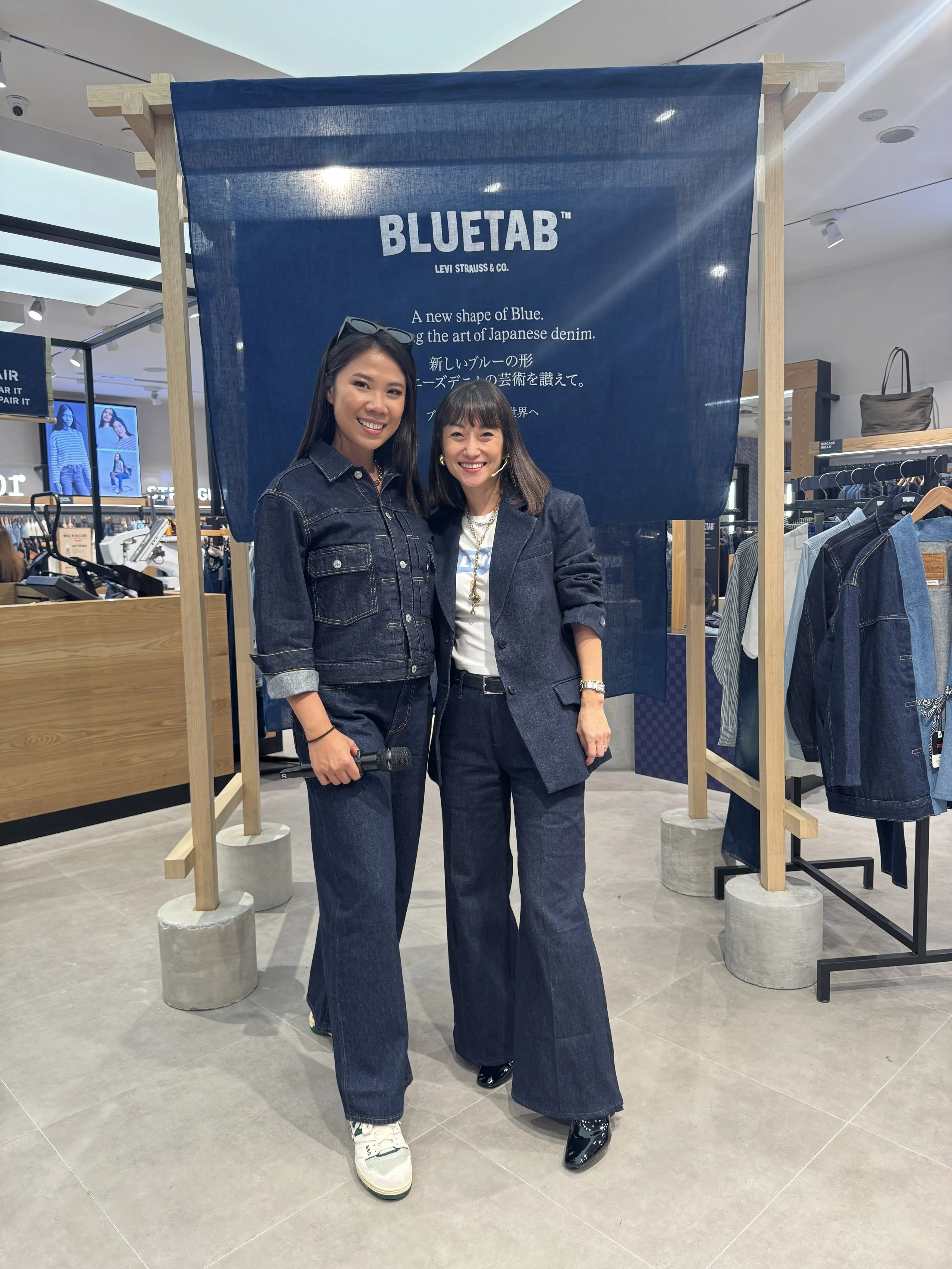 Two women in denim clothing standing in front of a blue fabric banner with text about Bluetab by Levi Strauss & Co., inside a clothing store. One woman holds a microphone, both are smiling.