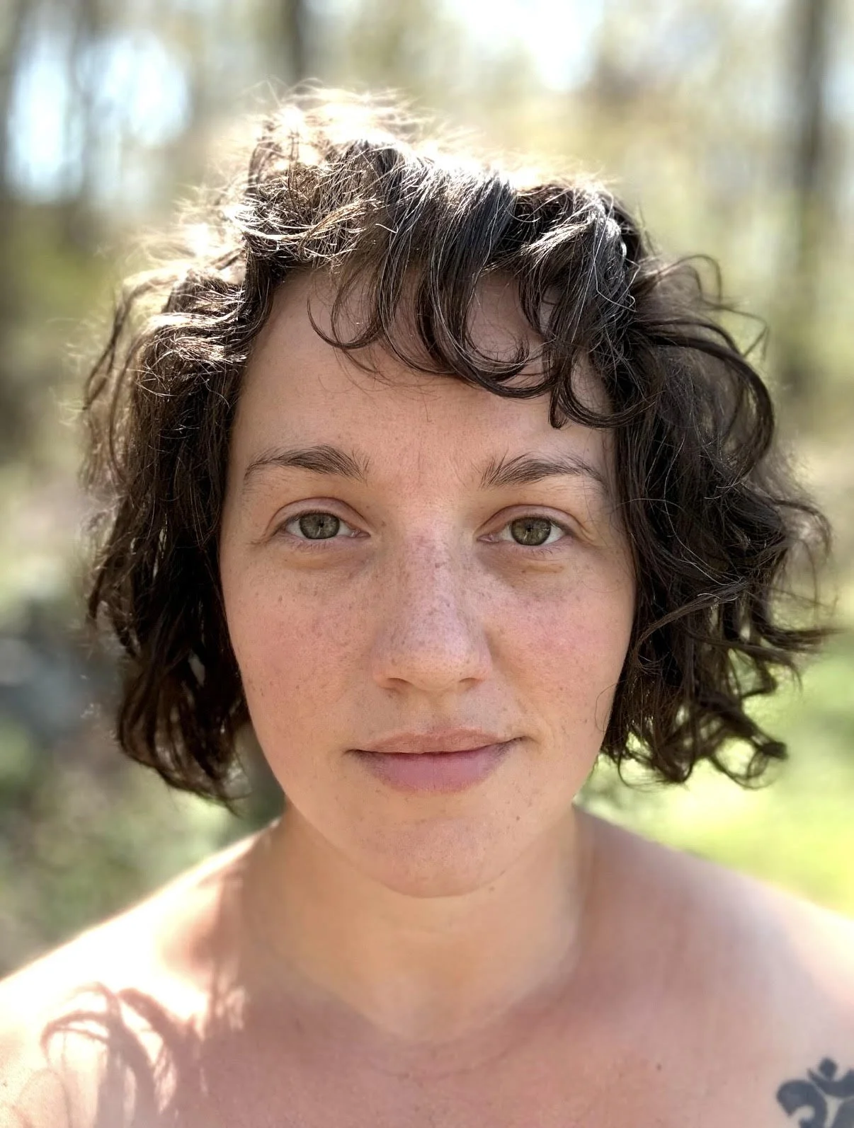 Close-up of a woman with short, curly dark hair and green eyes outdoors with a blurred natural background.