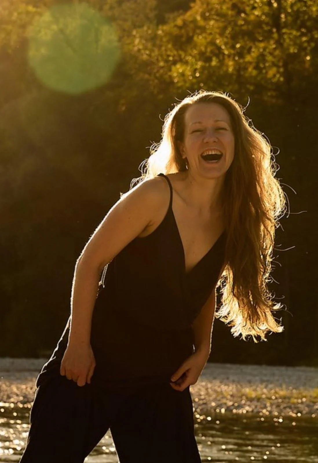 A woman with long red hair laughing outdoors during sunset, wearing a black dress.