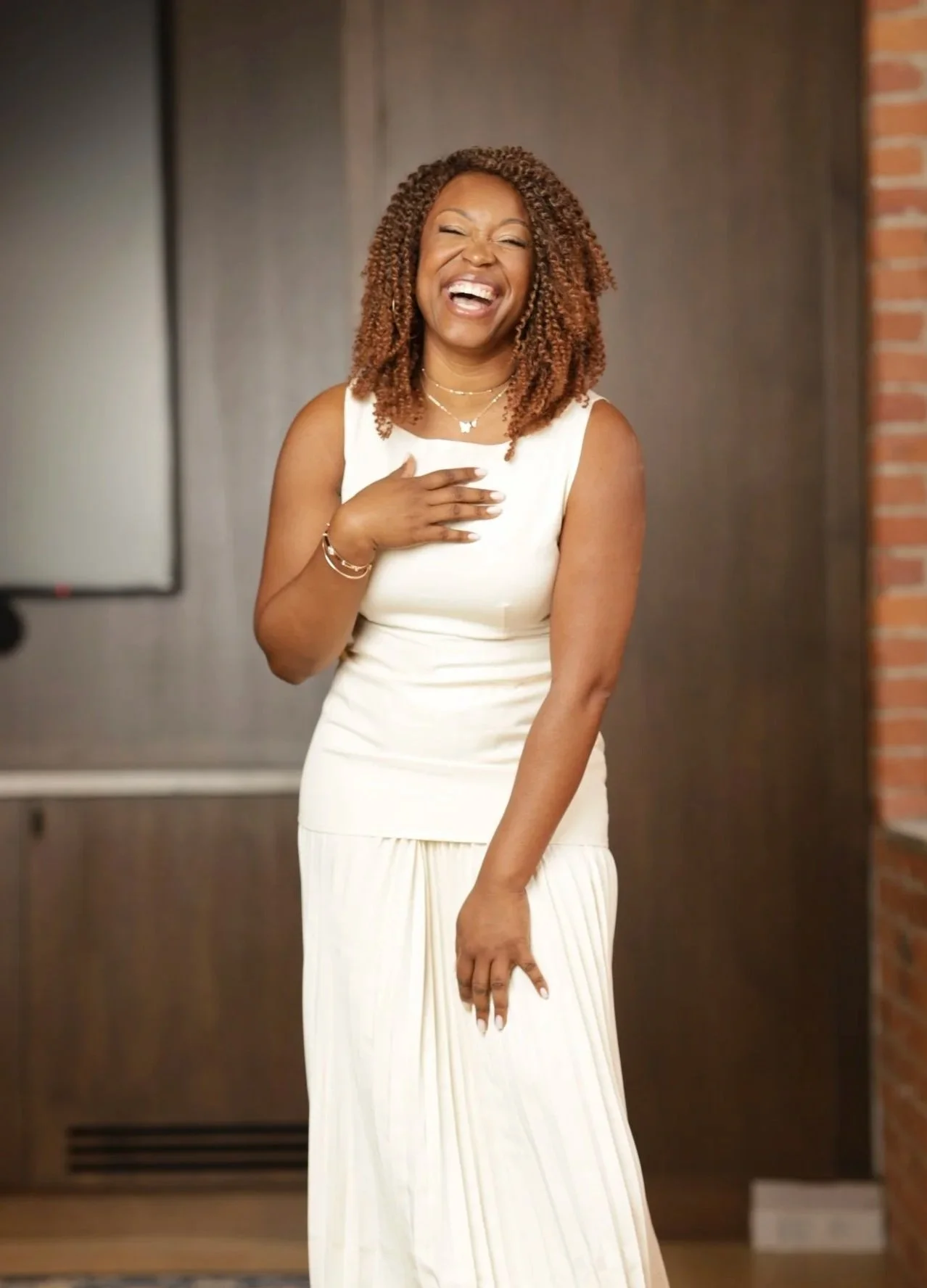 A woman in a sleeveless cream-colored dress laughing and covering her chest with one hand in front of a background with a dark wood panel and a brick wall.