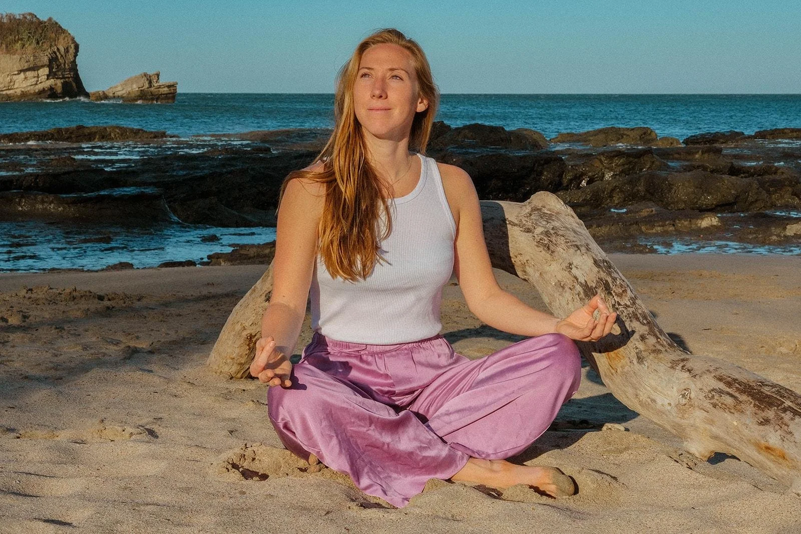 A woman in a white tank top and pink pants practicing yoga on a sandy beach, sitting cross-legged with eyes closed near a large piece of driftwood, with the ocean and rocks in the background.