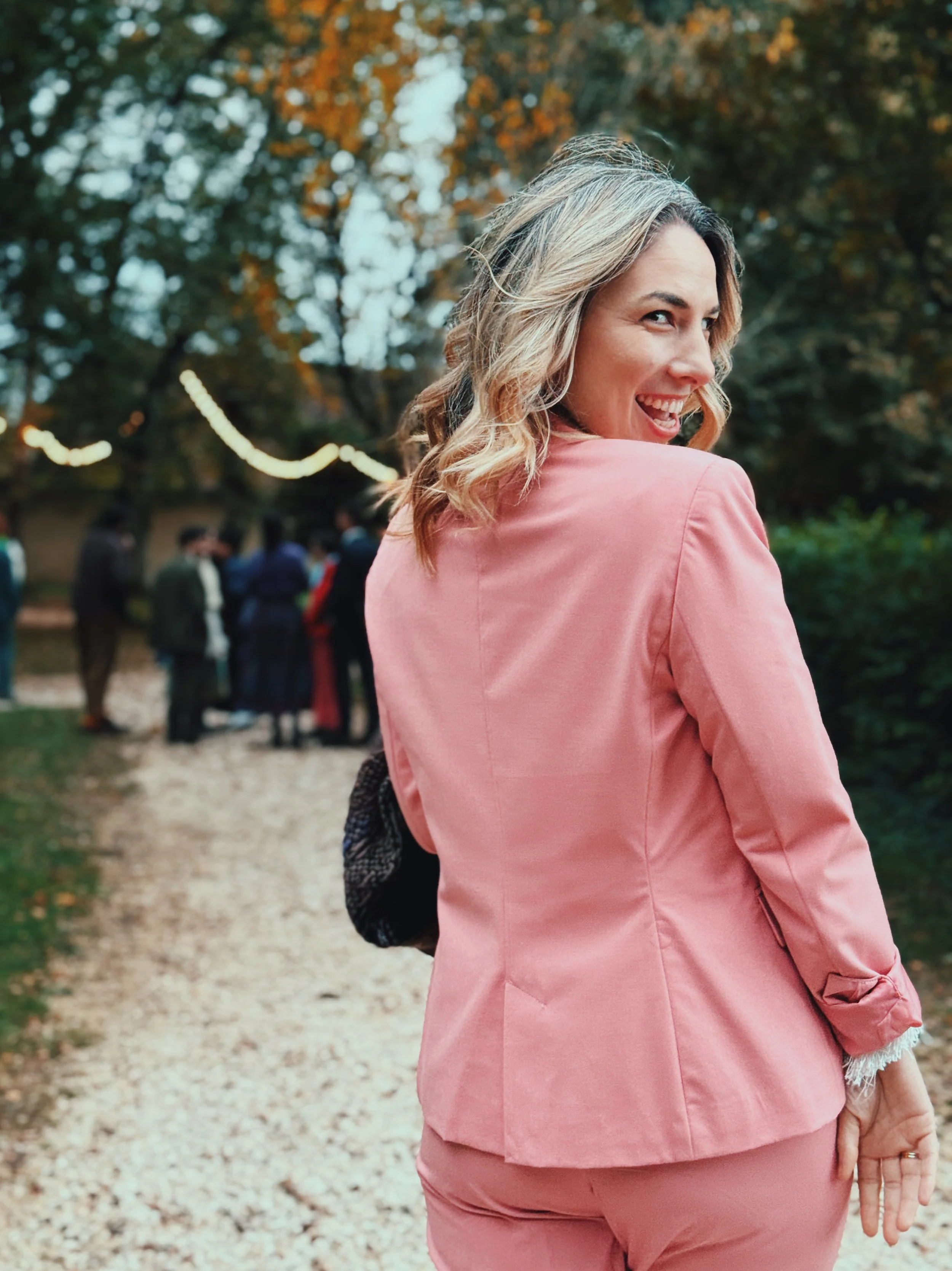 Woman with blonde hair smiling and turning back, wearing a pink suit, outdoors at a gathering during fall.