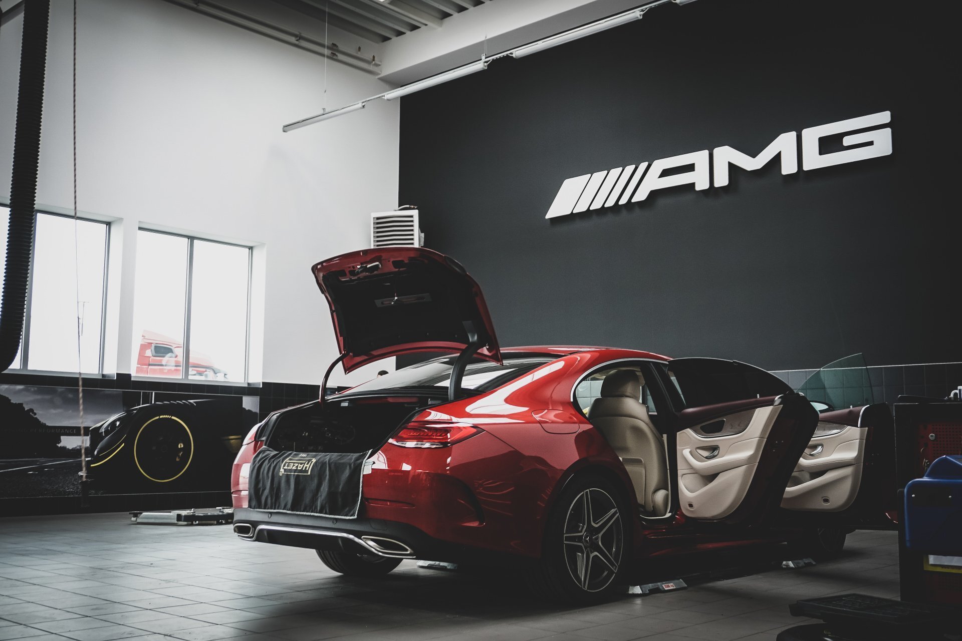 Interior of a luxury car showroom with a red sports car in the foreground, with its door open and trunk lifted, displaying a HRE Wheels cover. In the background, there is a black sports car, large windows letting in natural light, and a dark wall featuring the AMG logo.