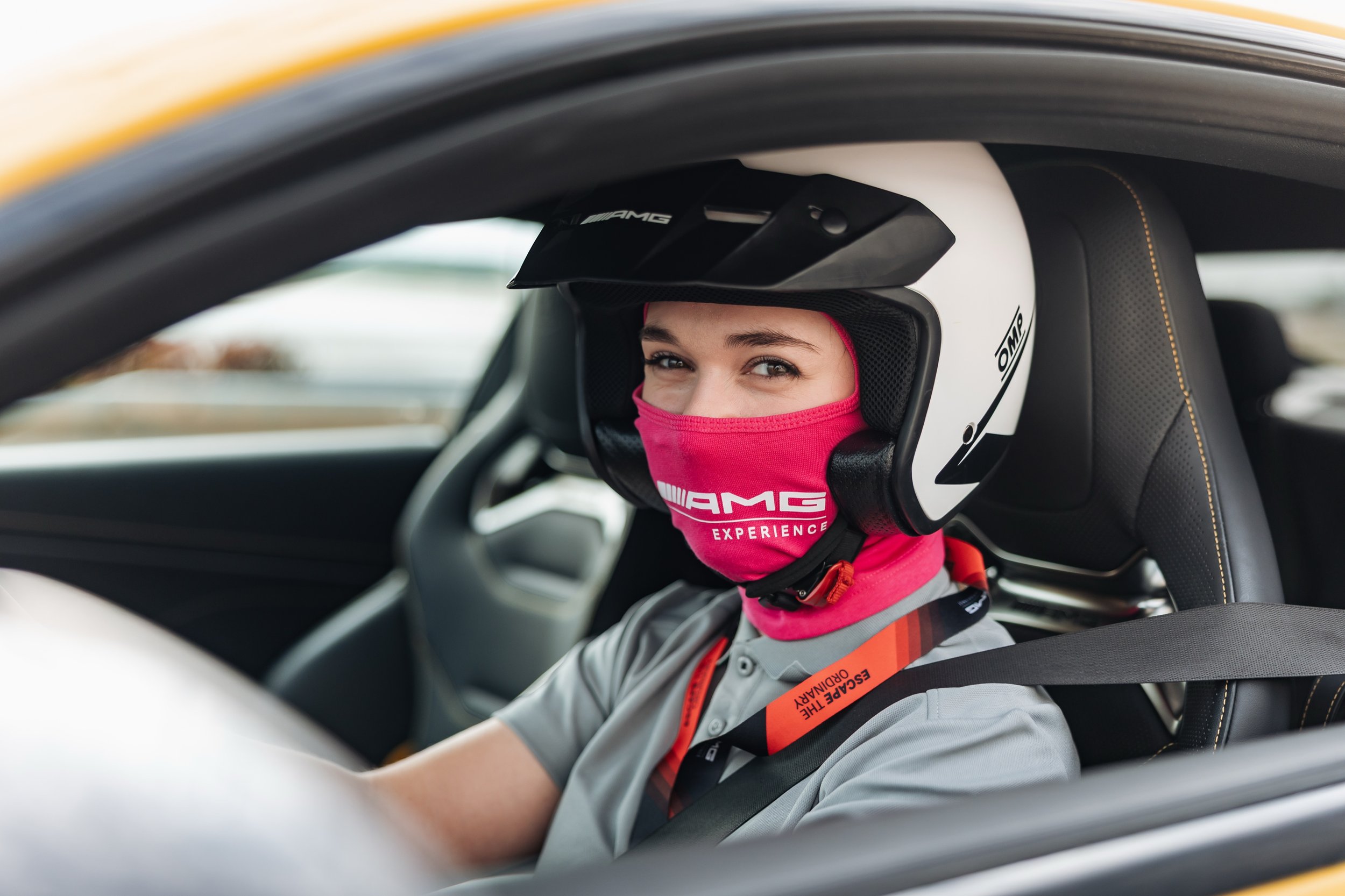 Kierowca wyścigowy (kobieta) w kasku i kominiarce z napisem »AMG EXPERIENCE« zapięty pasami w sportowym fotelu.