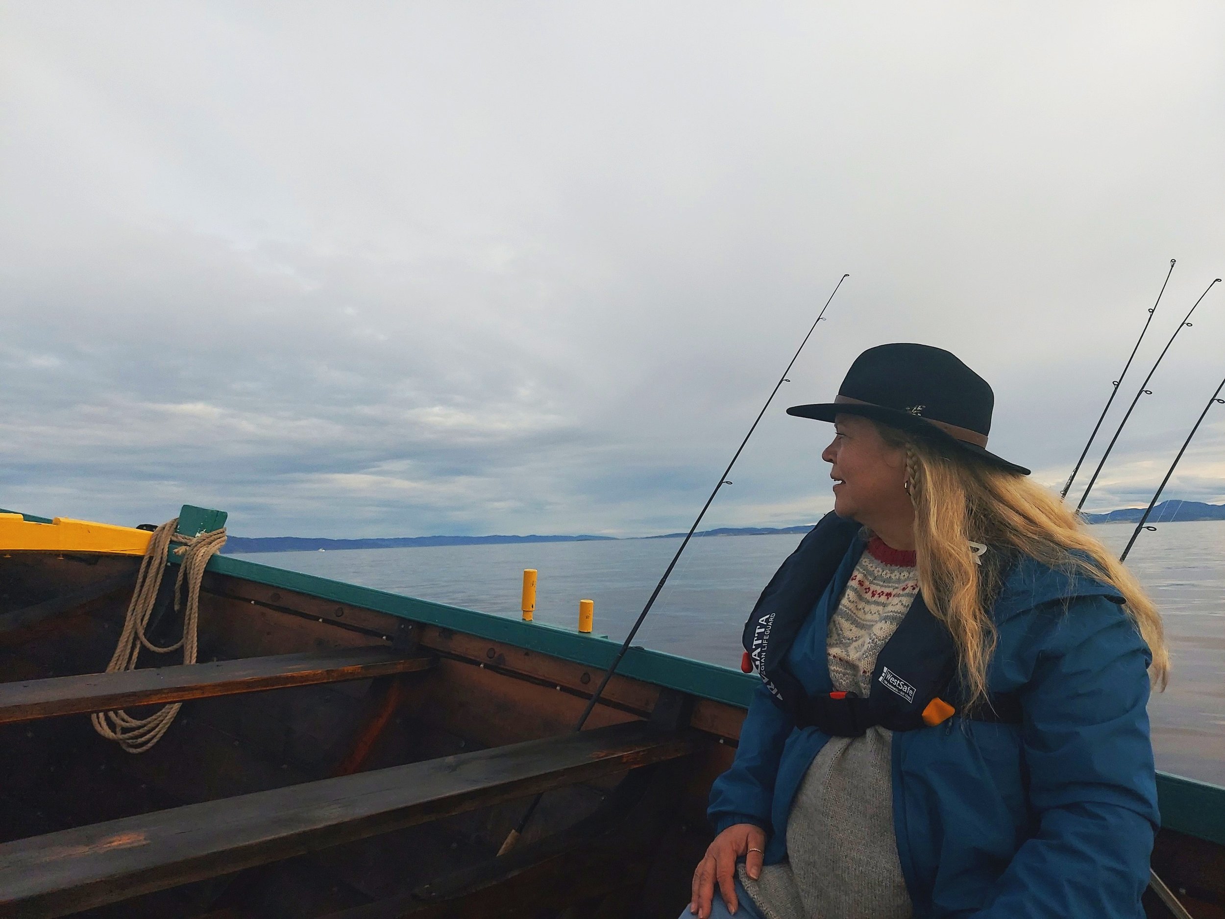 Kvinne i båt med fiskeutstyr, iført hatt og flytevest, ser på horisonten.