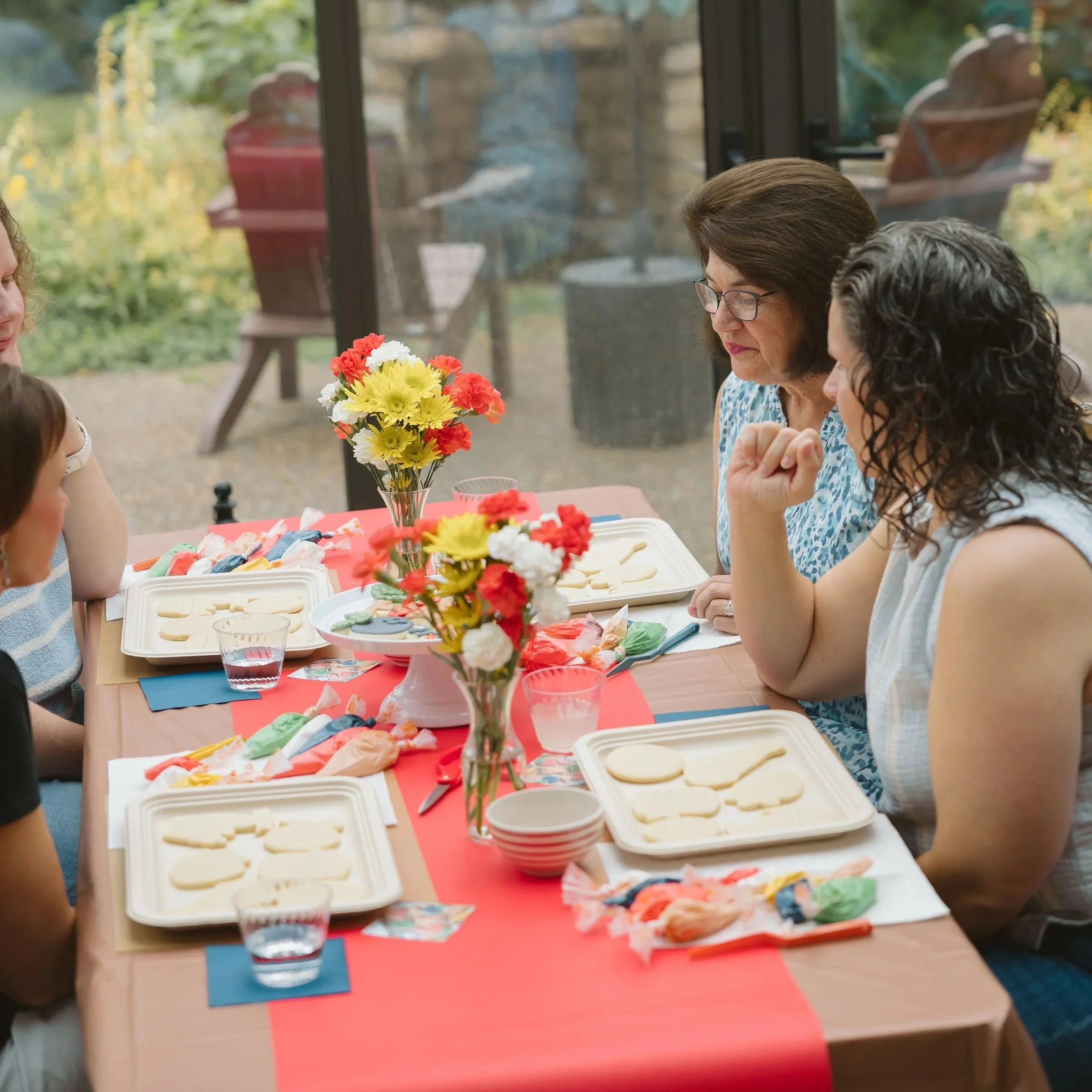 A family gathered around a dining table with decorated cookies, juice, colorful napkins, and a bouquet of flowers, inside next to a large window overlooking a garden.