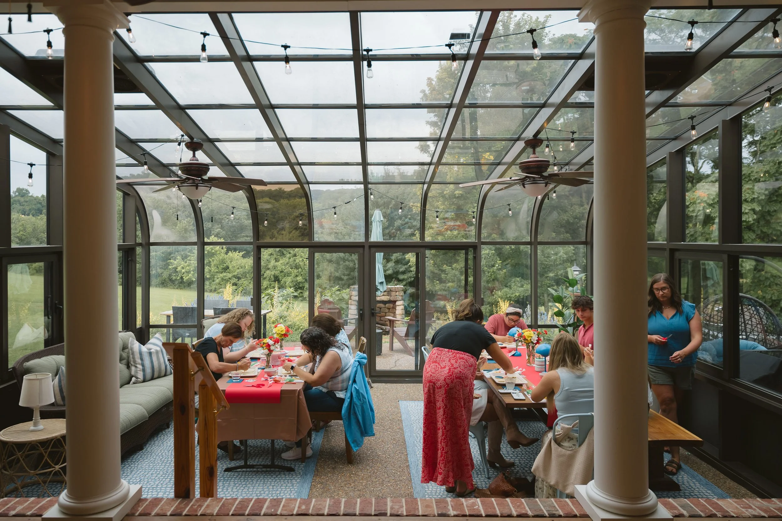 People gathered at a long table inside a glass-enclosed sunroom with ceiling fans, engaged in a craft activity, with floral centerpieces and outdoor scenery visible through the windows.