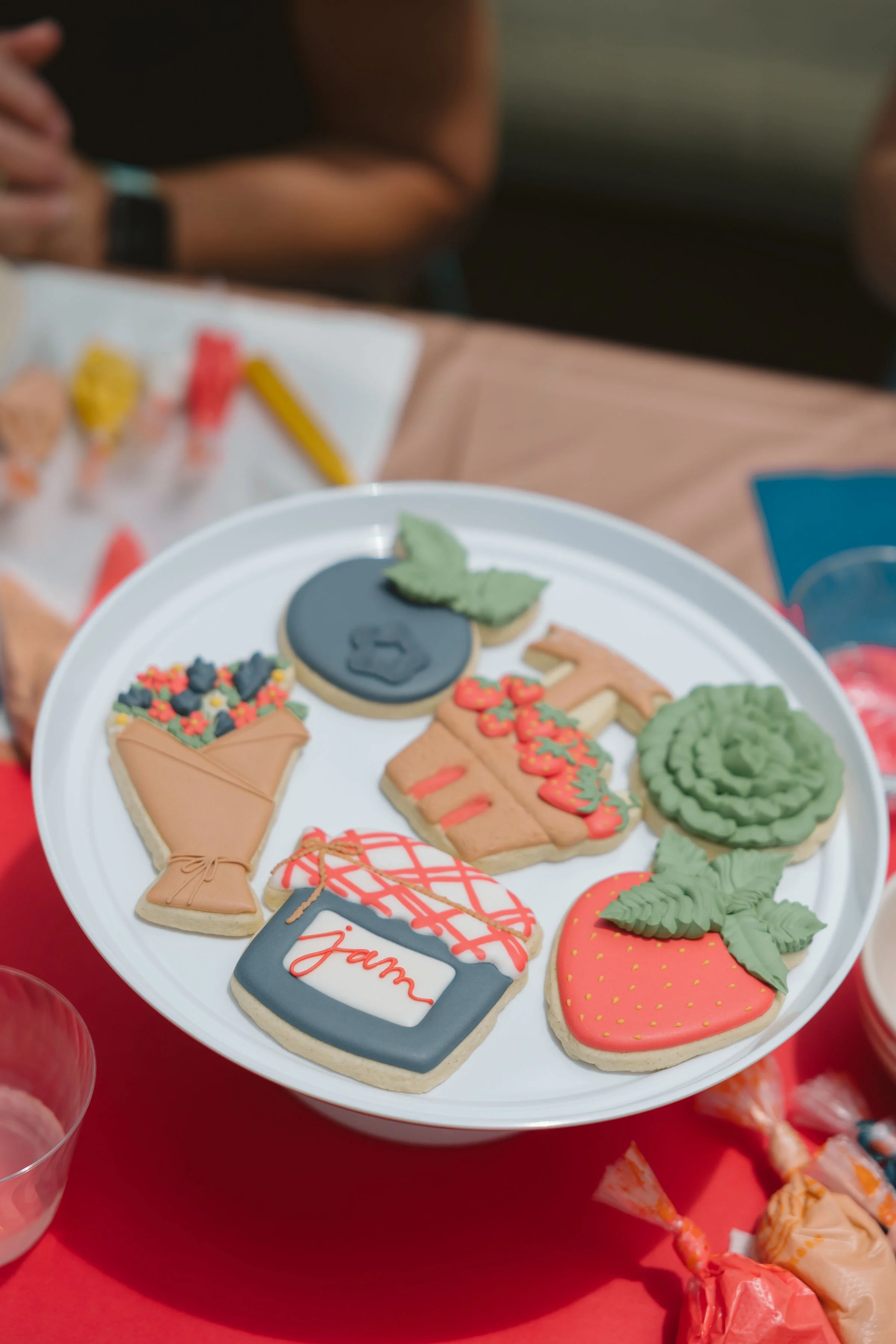 Assorted decorated cookies on a white tray, including a strawberry, a jar of jam, a rose, a blackberry tart, a hot pepper, and a blueberry, with a person partially visible in the background.