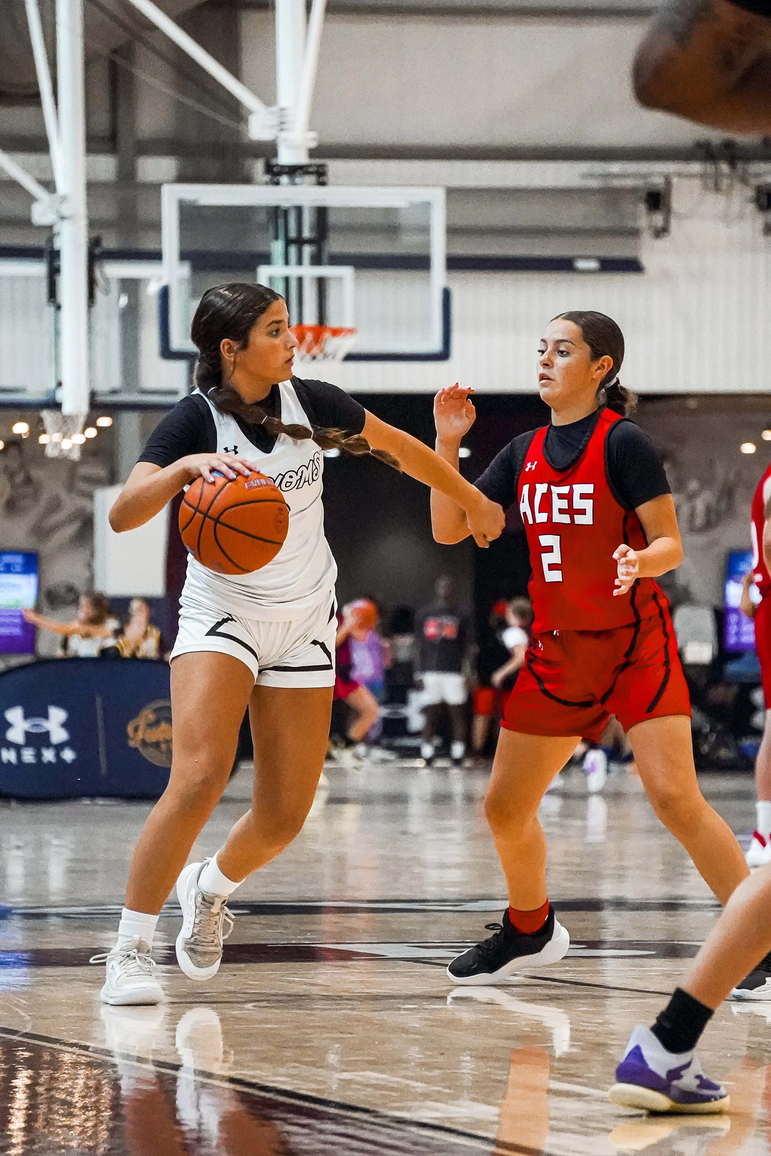 Two female basketball players compete for possession of the ball during a game inside a gymnasium, with a basketball hoop in the background.