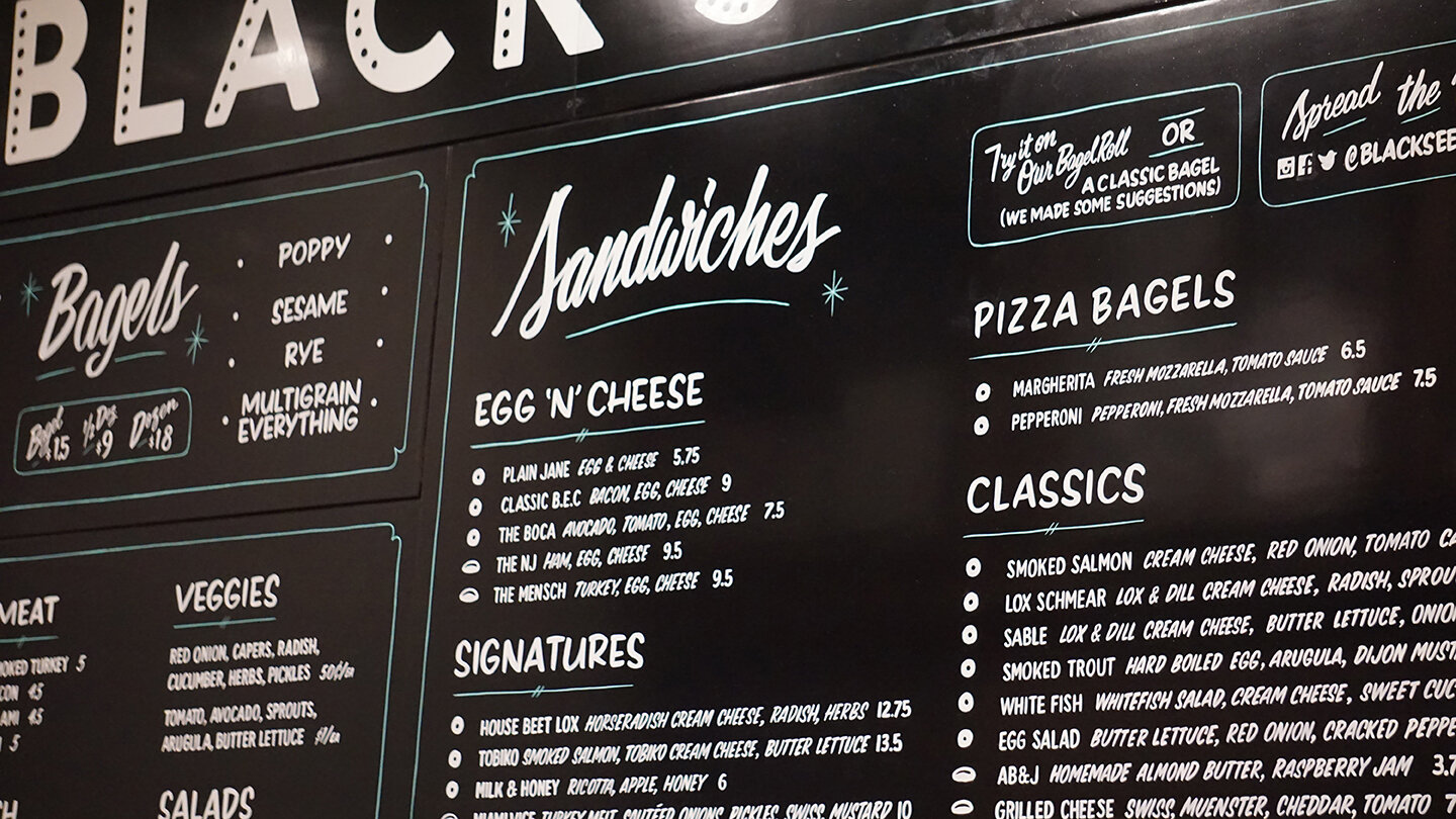  Close-up of a handpainted menu board for Black Seed Bagel with white lettering on a black backdrop  