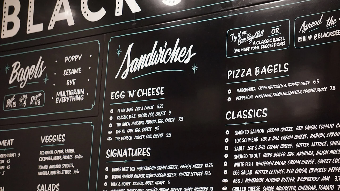  Close-up of a handpainted menu board for Black Seed Bagel with white lettering on a black backdrop  