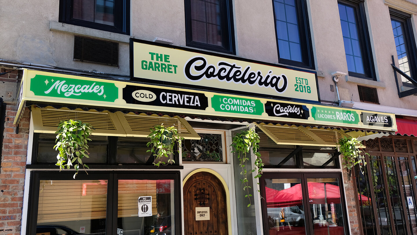  Yellow storefront sign reading “Cocteleria” in handpainted black casual script lettering, with handpainted green and black bodega-style sign elements describing the drink options inside 