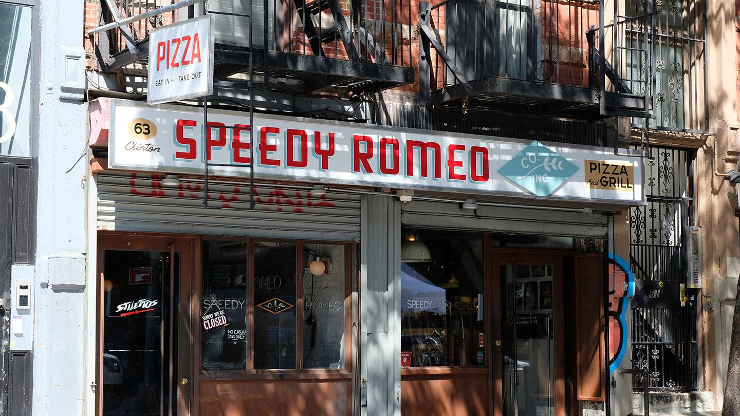  Storefront sign reading “Speedy Romeo” in red block handpainted lettering on white backdrop for a pizza restaurant 