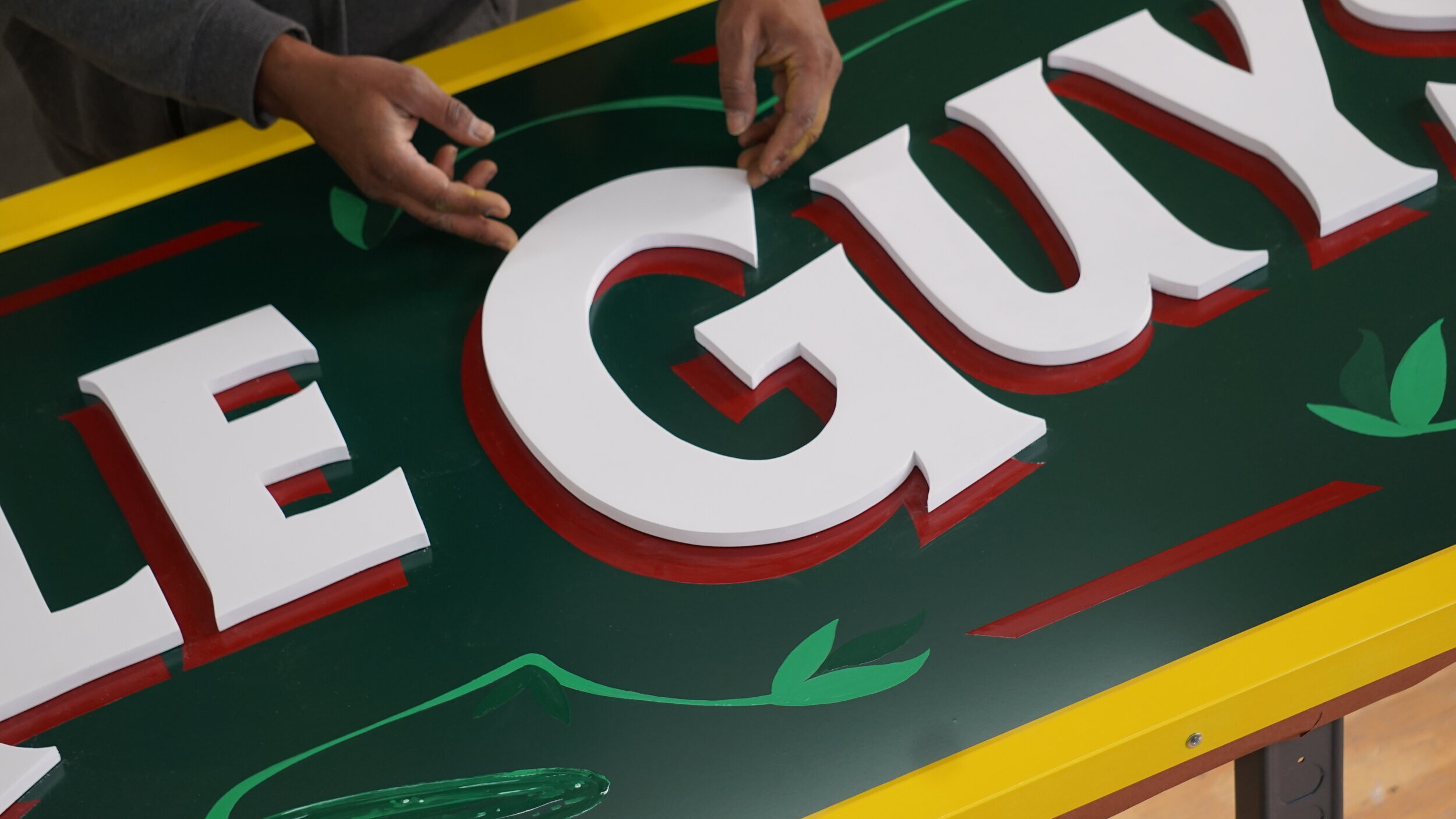  Close-up of the sign for Pickle Guys in Essex Market being fabricated: white dimensional cut letters are being mounted onto a dark green sign with red, green, and yellow decorative elements 