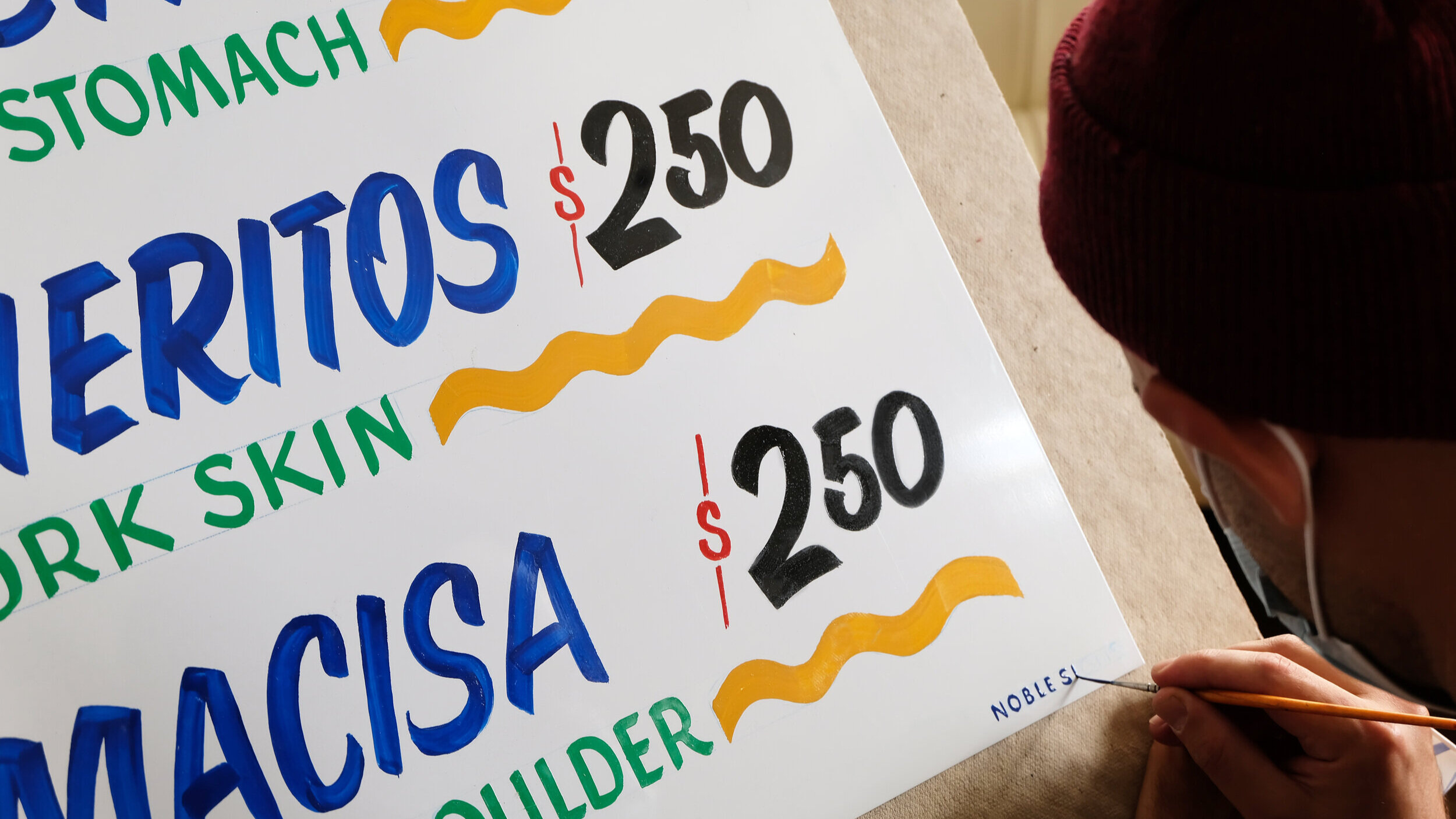  A close-uo of a restaurant menu being handpainted on paper in a casual signpainter’s lettering style 
