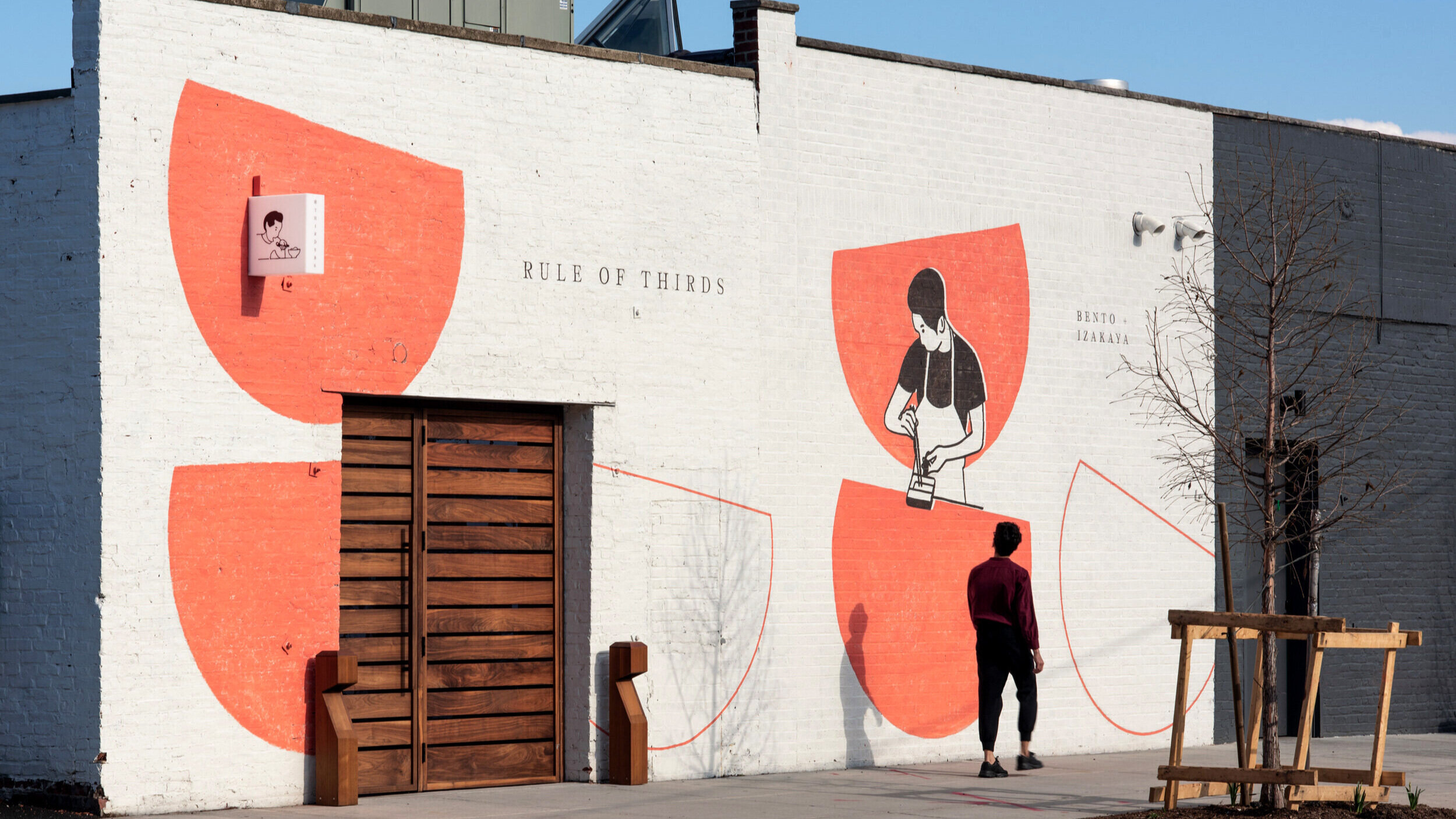  A white storefront exterior with a handpainted mural of bowls and a person cooking in orange and black paint, handpainted lettering reading “Rule of Thirds” in serif lettering, as well as a small lightbox blade sign of a person eating 