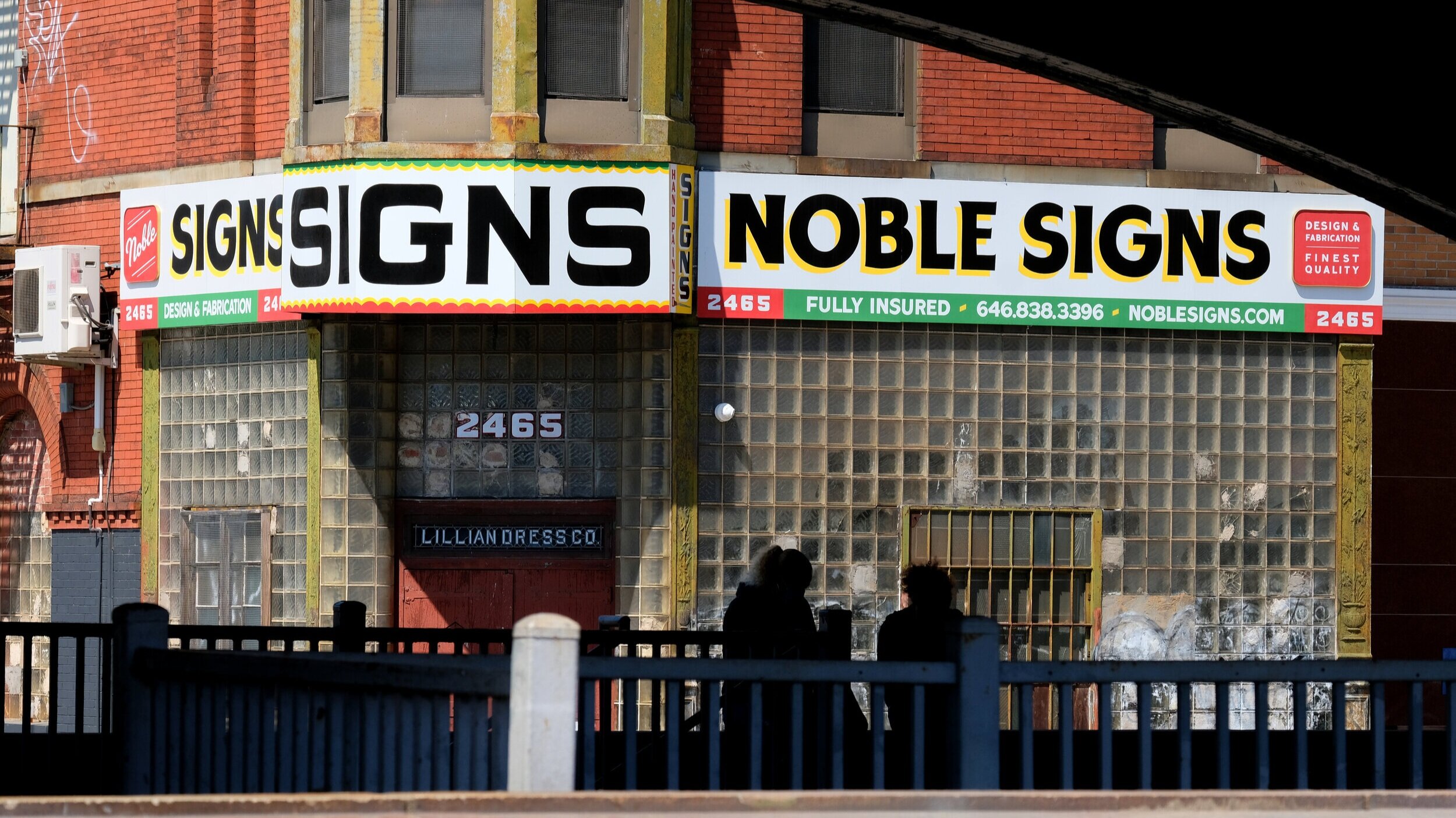  The Noble Signs studio sign, inspired by classic signage of NY in decades past, consisting of handpainted black block lettering with a yellow drop shadow on a white backdrop, with red and green elements containing various information along the botto