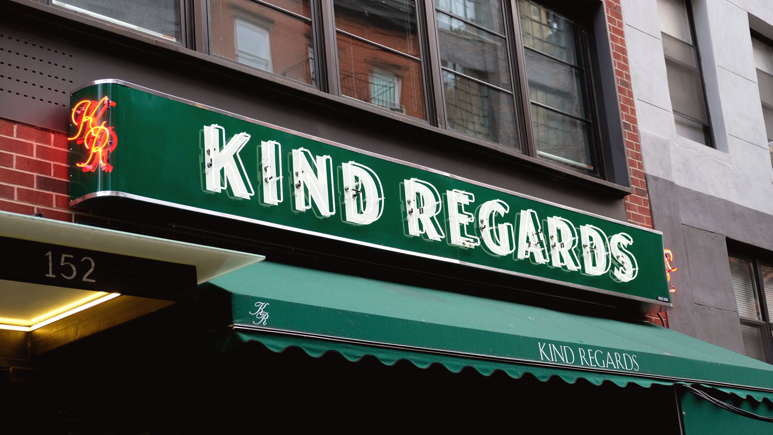  Handpainted white block lettering with overlaying white neon tubing, reading “Kind Regards” on a dark green storefront sign, as well as a red neon reading “KR” in decorative lettering mounted on the side edge 