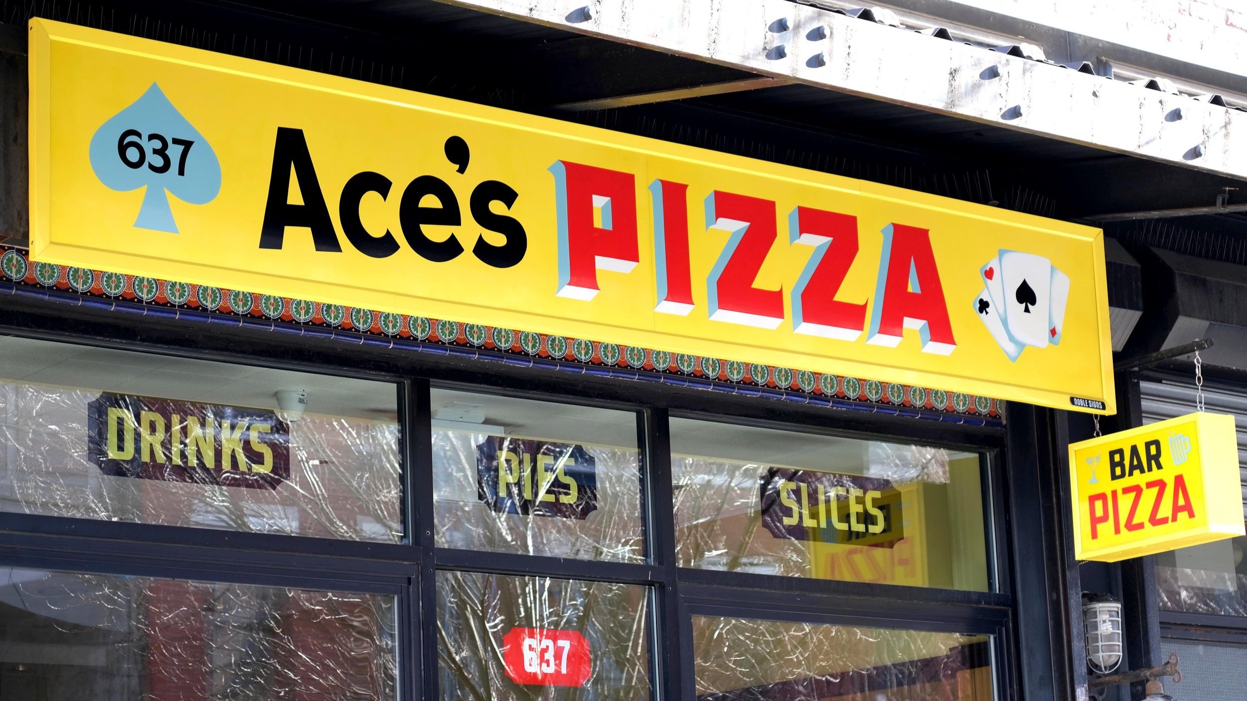  Yellow storefront sign reading “Ace’s Pizza” in black and red handpainted block lettering, with paintings of a spade and a hand of cards on the sides 