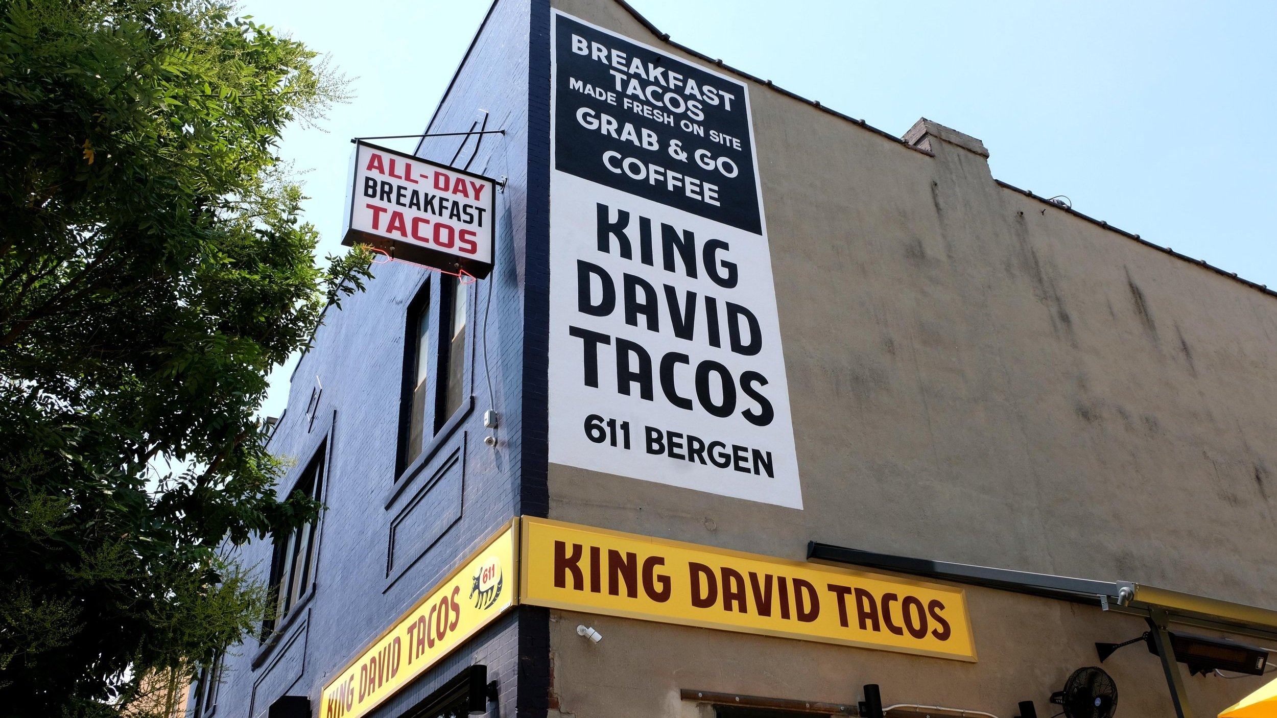  A sign handpainted onto an exterior wall reading “King David’s Tacos” in block lettering and black and white paint, a yellow storefront sign reading the same in red, and a blade sign reading “All Day Breakfast Tacos” 