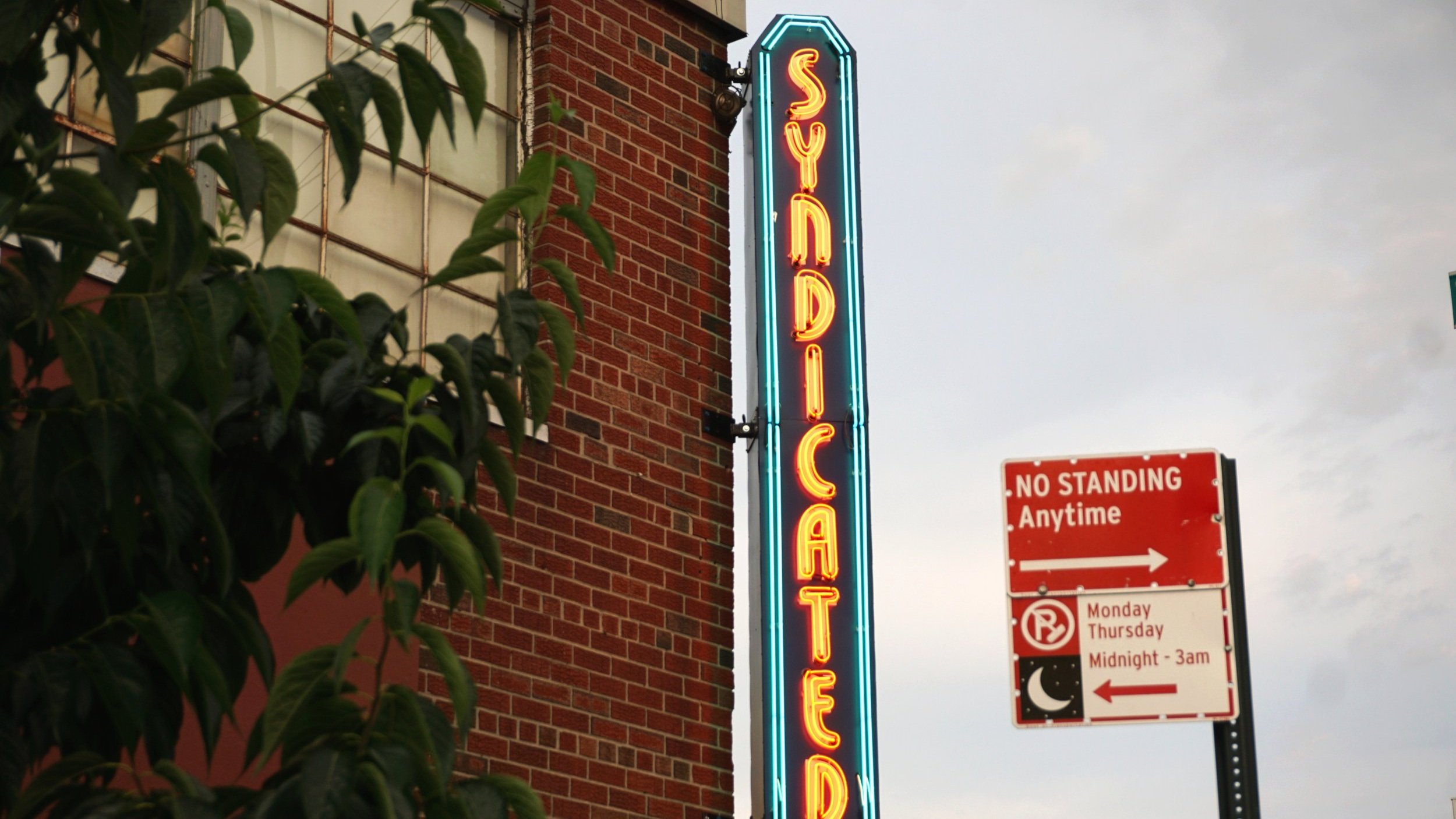  A vertical neon blade sign projecting off a building corner reading “Syndicated” in red neon with an aqua neon border 