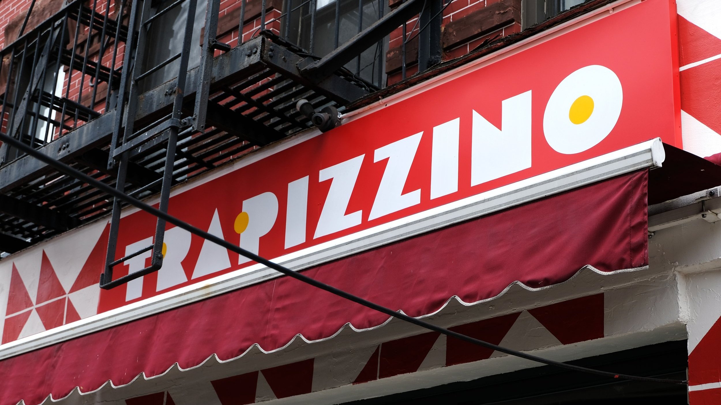  A red storefront sign with white handpainted decorative geometric lettering reading “Trapizzino” 