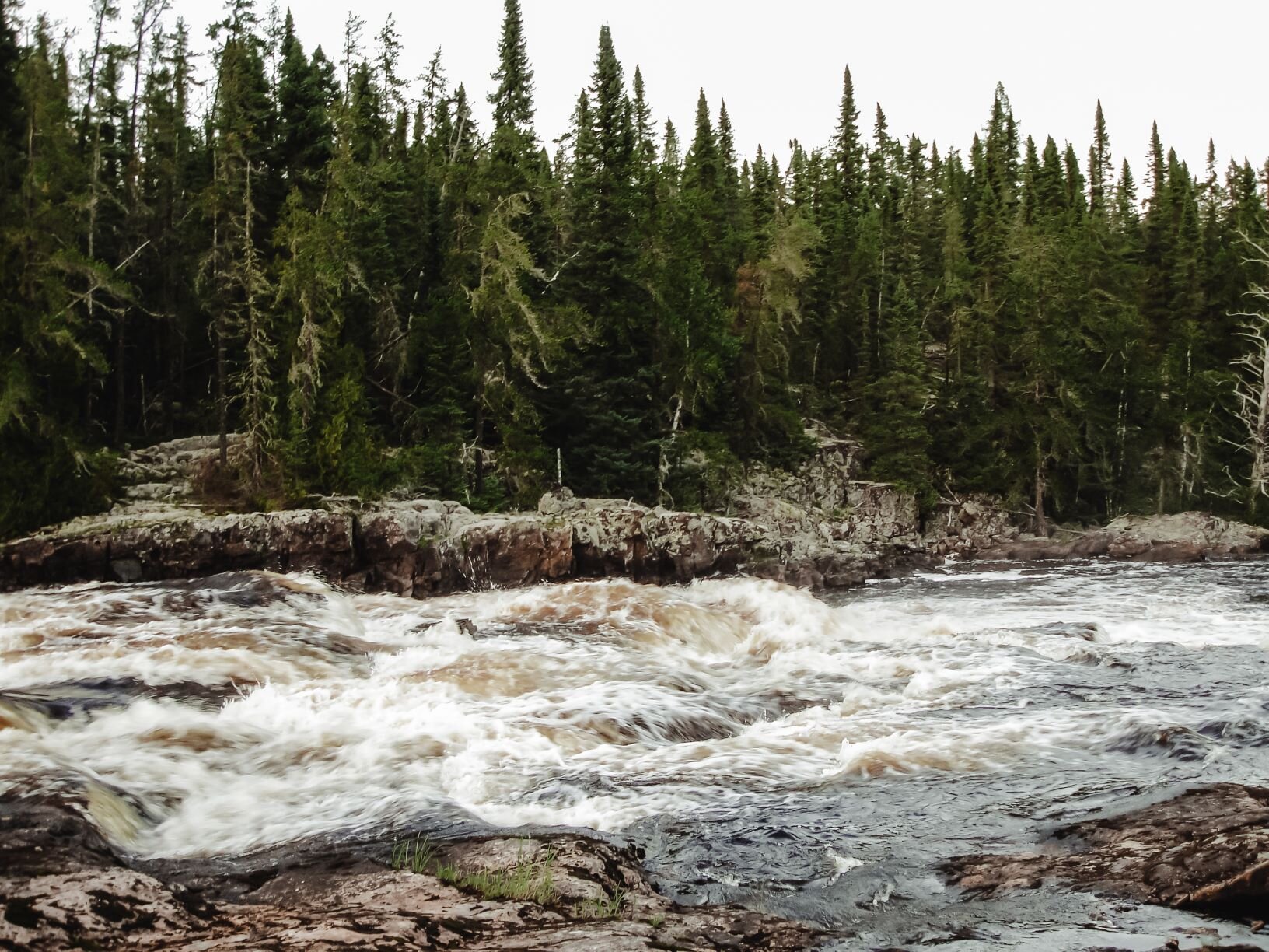 A photo of a rushing river. The shorelines on either side are made of rock, and the far shore is covered in evergreen trees.
