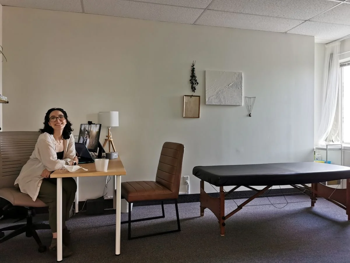 A wide-angle photo of a room with white walls with some art, a low massage table, a simple desk, with a comfortable chair with no armrests on one side, and Arlie sitting in an office chair and smiling on the other side.