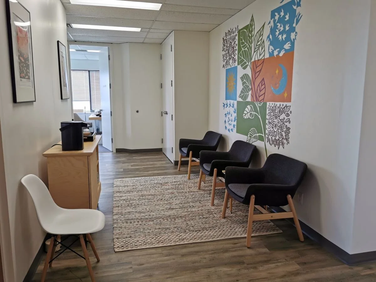 A photo of a room with a mural on one wall with three grey upholstered chairs below it, a grey rug on the floor, and a sideboard with a water dispenser and cups along the opposite wall.