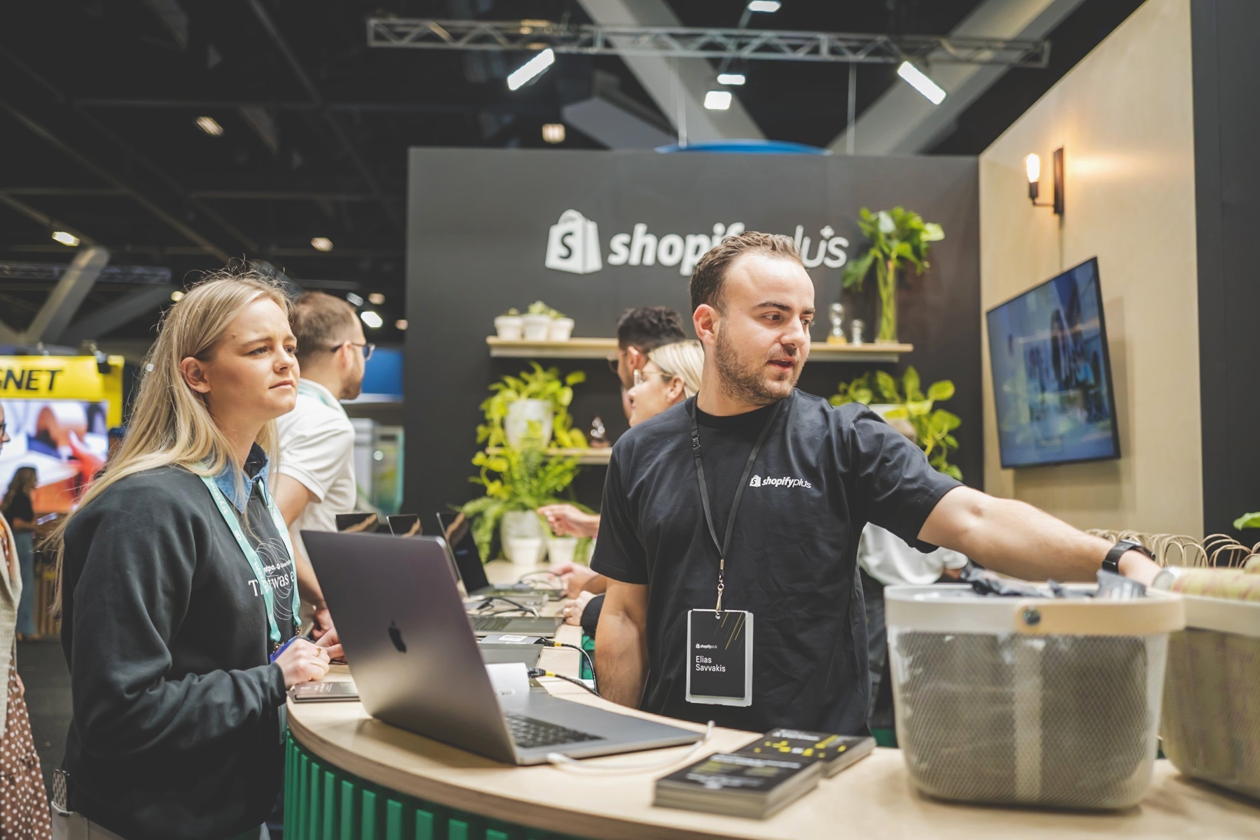 A man behind a counter at a booth with a laptop and multiple onlookers, at the Shopify Plus exhibit, with plants and a screen displaying content in the background.
