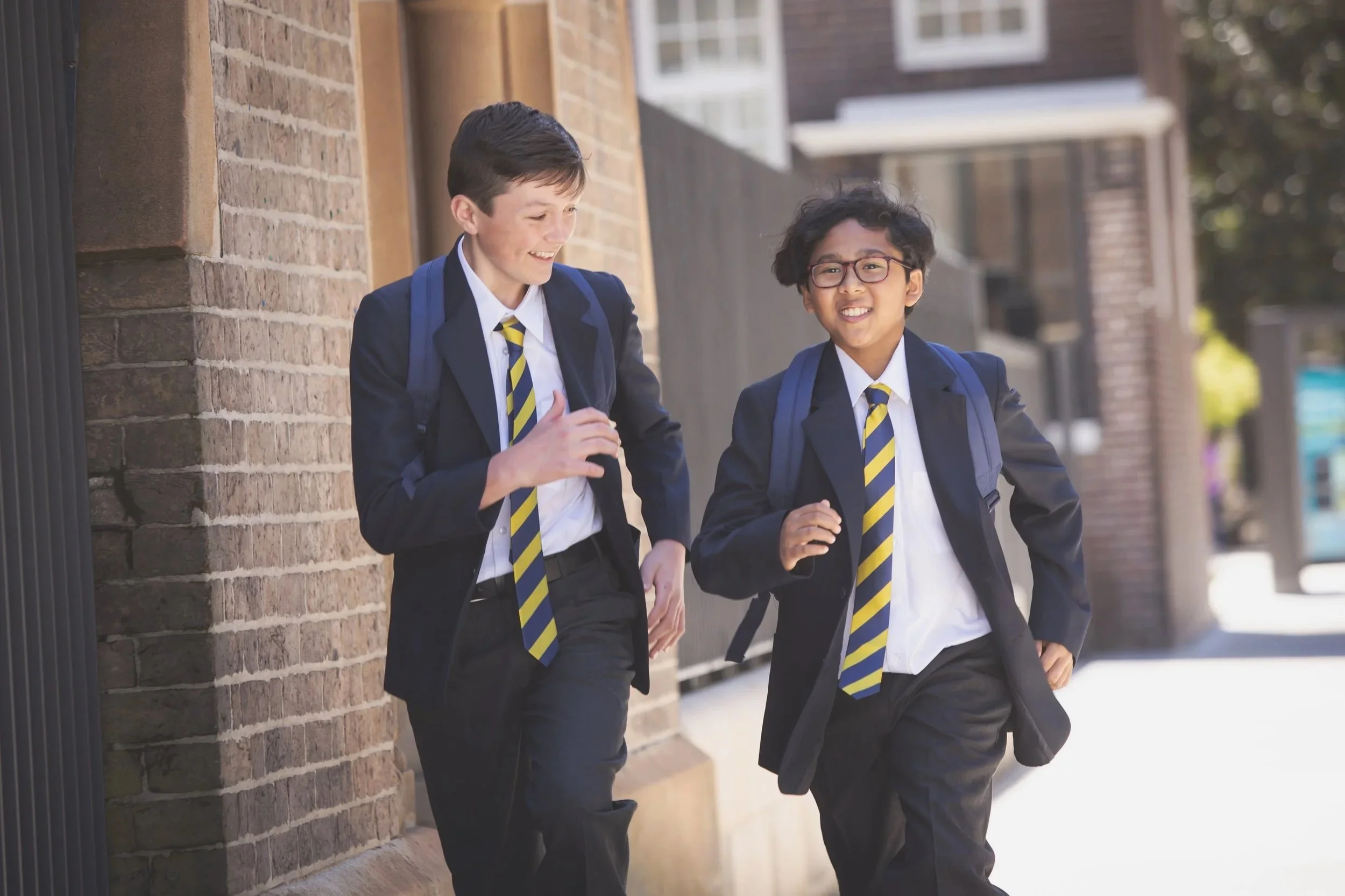 Two schoolboys in uniform walking and talking outside a brick building, smiling, wearing blazers and striped ties, carrying backpacks.