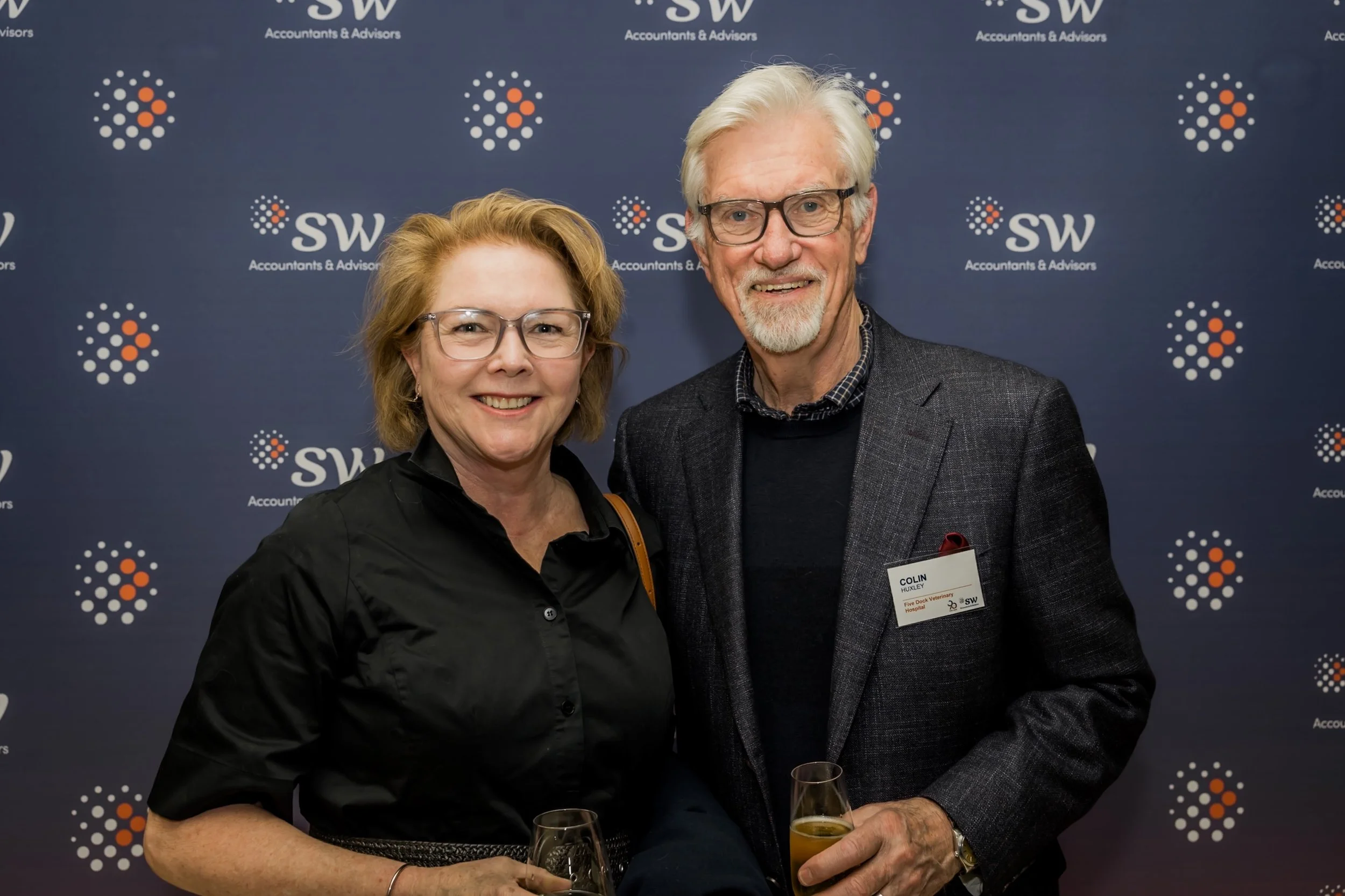 Two smiling people, a woman and a man, standing together at an event with a backdrop that has the logo 'SW Accountants & Advisors'. The woman has shoulder-length blonde hair, wears glasses, and a black shirt. The man has white hair, a gray beard, gla