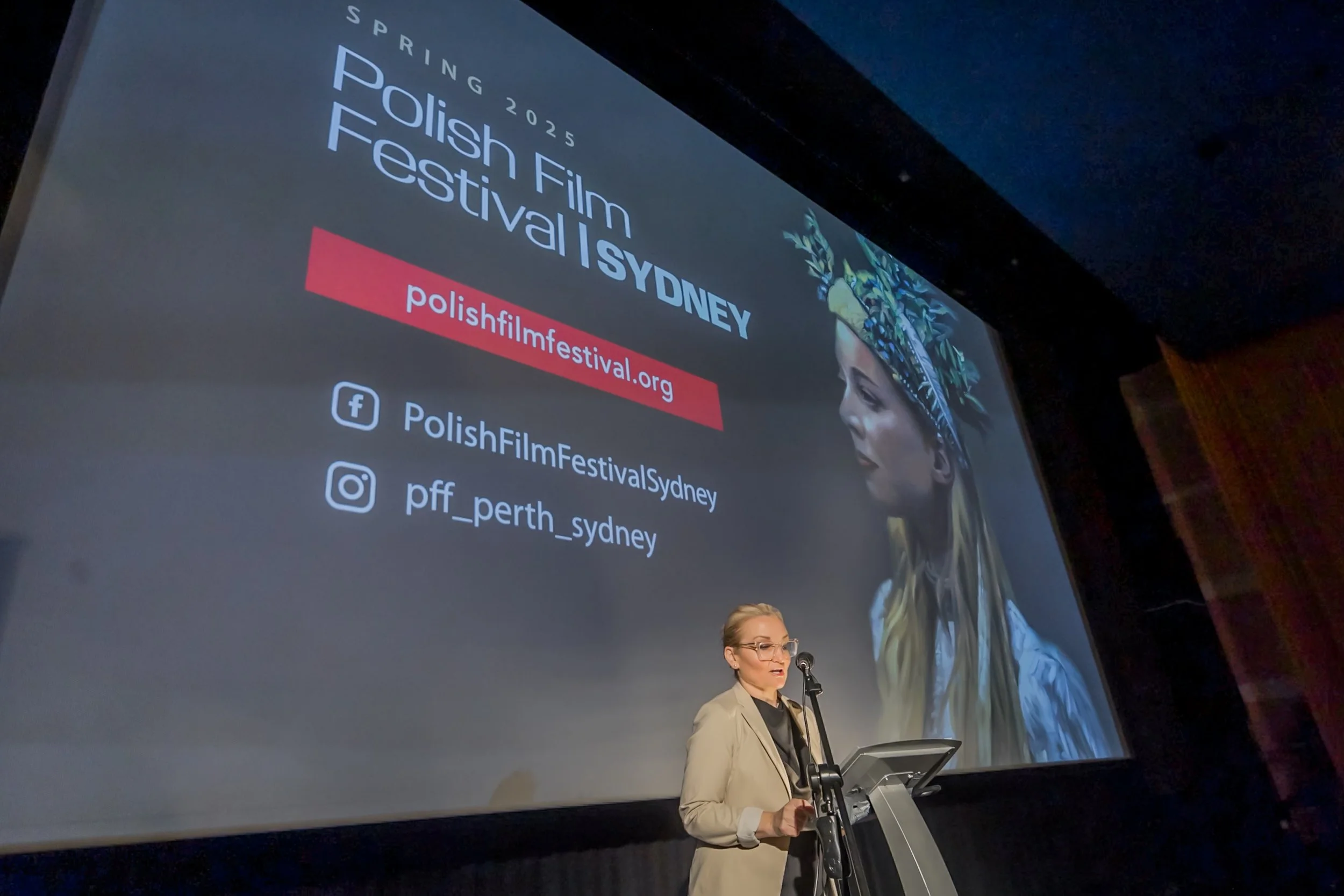 Woman speaking at a podium during the Polish Film Festival Sydney event, with a large screen displaying event information behind her. The screen shows the festival's website, Facebook, and Instagram handles, and features an image of a woman wearing a