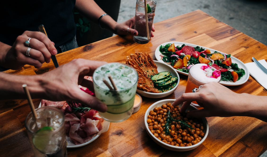People sharing a meal at a wooden table with salads, chickpeas, cold drinks, and a green cocktail with cinnamon sticks.