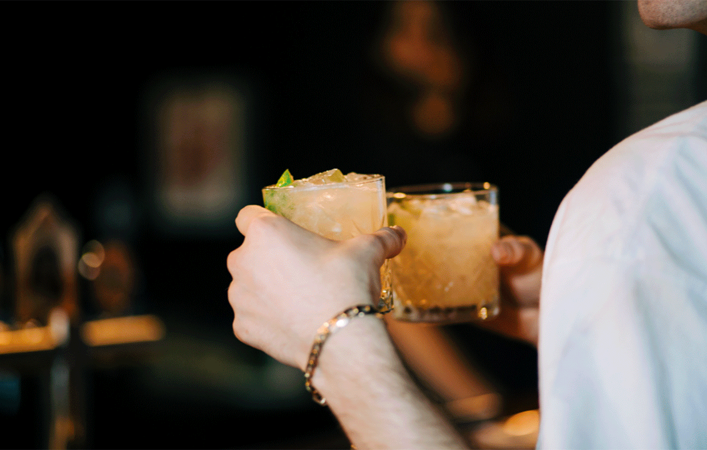 Person holding two glasses of cocktails with lime garnish, clinking them together in a toast.