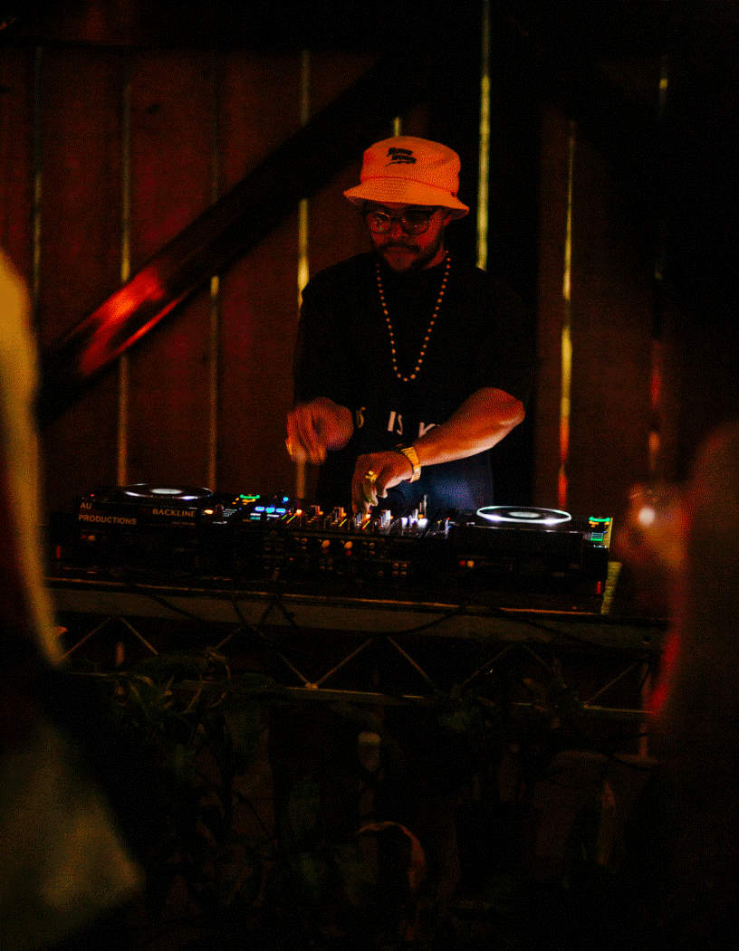 A DJ wearing a beige bucket hat, glasses, and a beaded necklace, standing behind a DJ controller in a dimly lit setting.