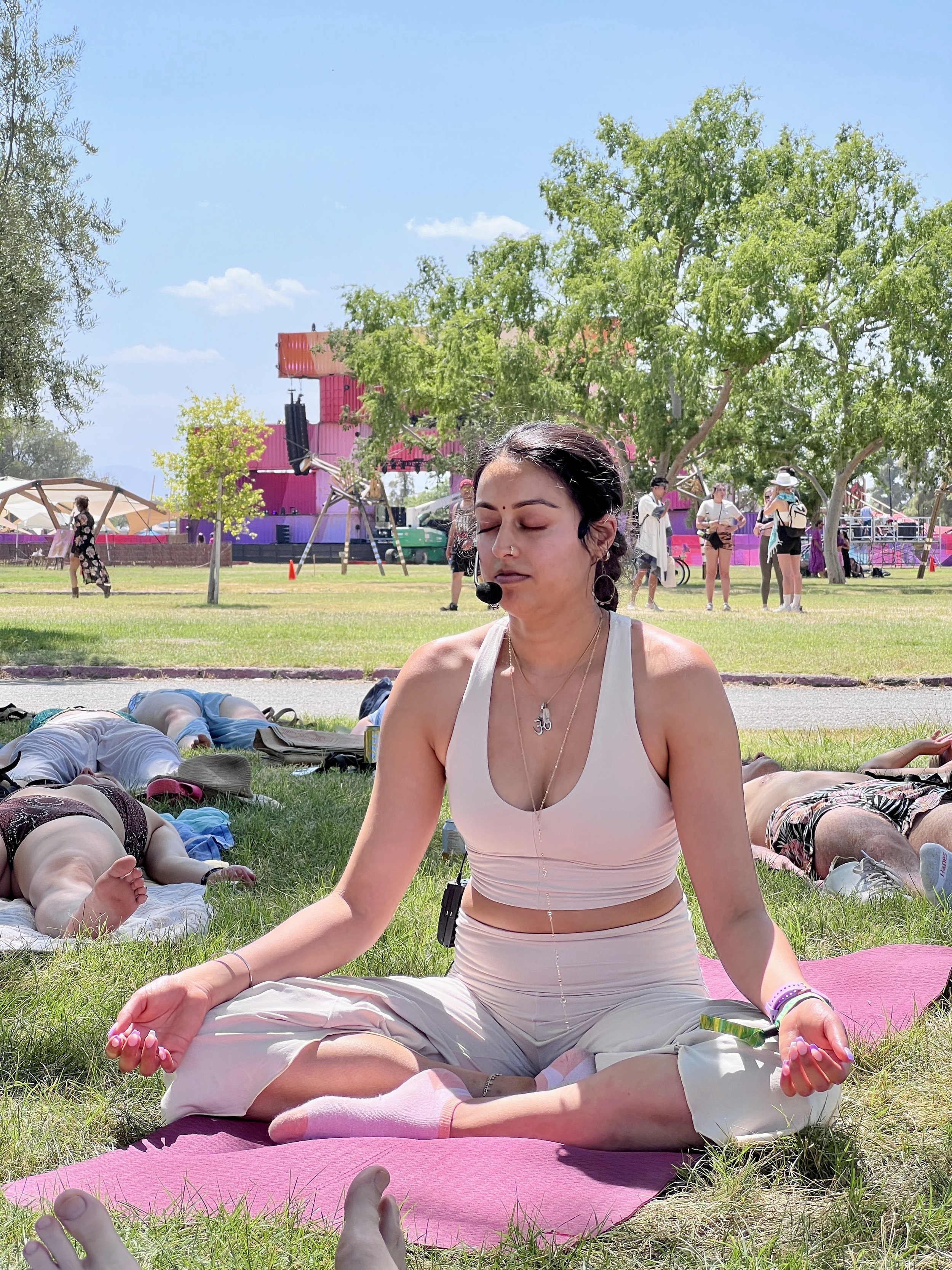 A woman practicing yoga outdoors on a pink mat, seated in a cross-legged meditation pose with eyes closed, surrounded by other people lying on mats in a park, with trees and a colorful playground structure in the background.