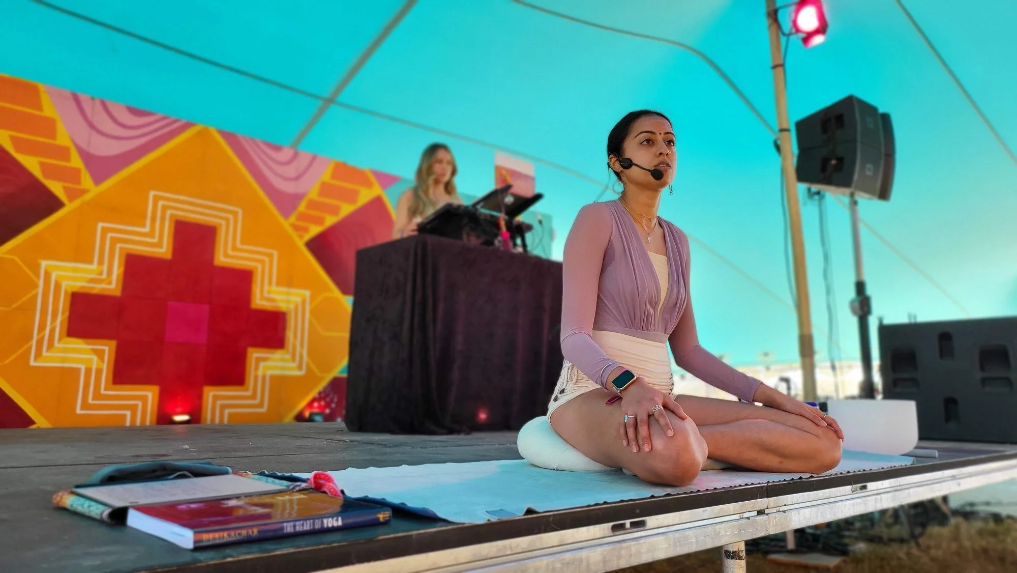 A woman practicing yoga and meditation outdoors under a turquoise canopy, with a DJ in the background and colorful geometric artwork on the wall.