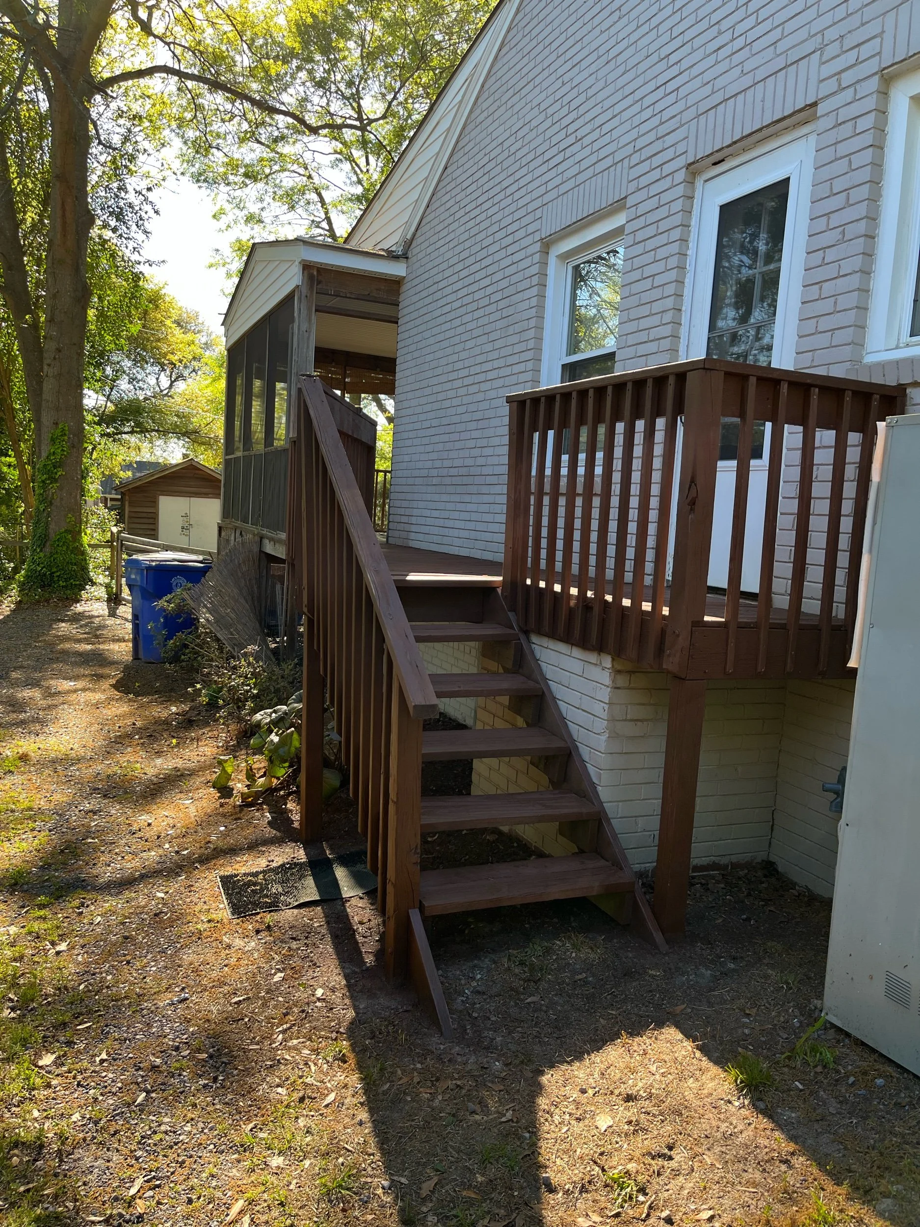 Backyard view of a house with a wooden staircase leading to a small deck and a screened porch. Trees surround the area, and there are some blue recycling bins to the left.