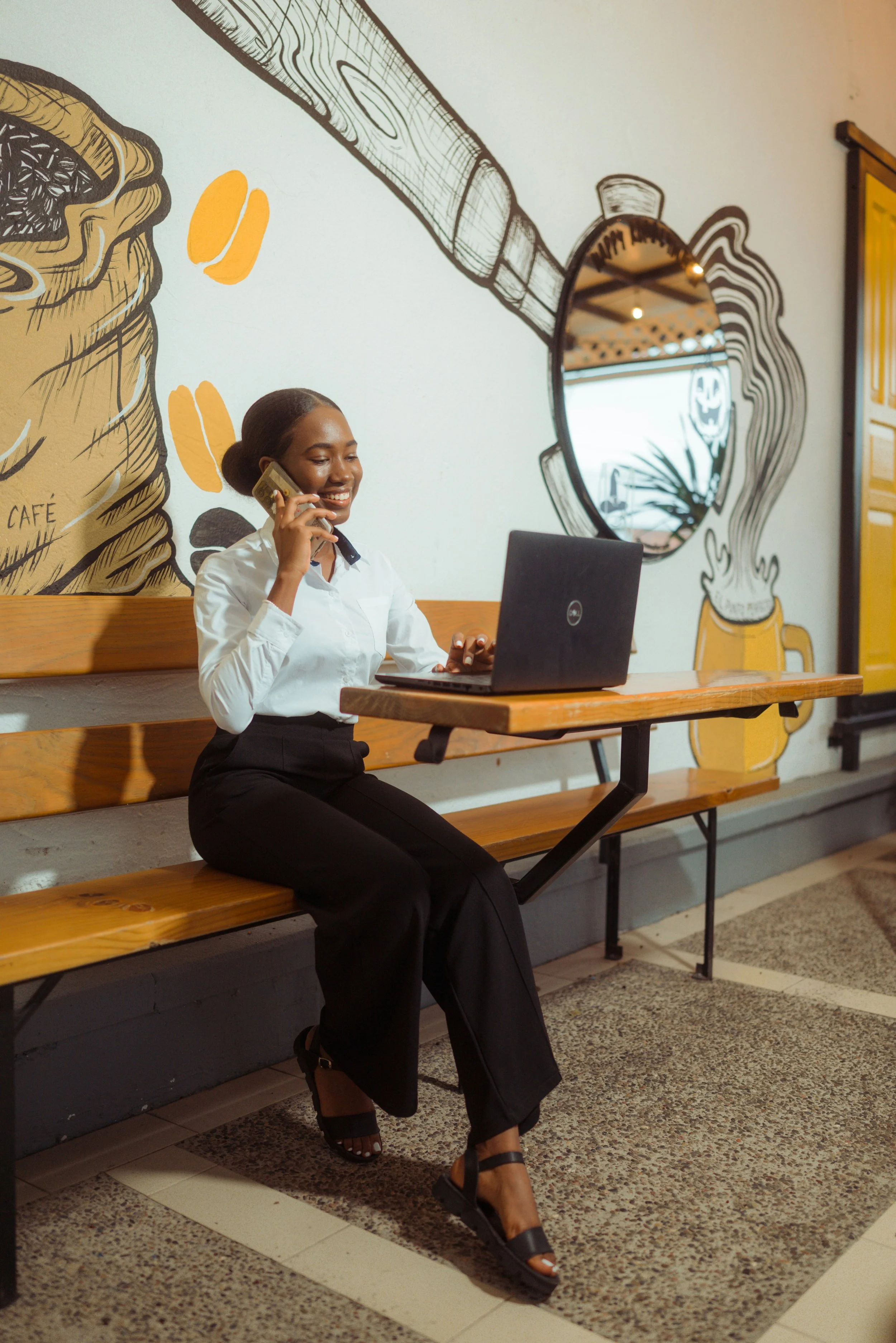 A woman sitting on a wooden bench at a cafe, talking on her cellphone, with a laptop open in front of her, smiling. The wall behind her has a black and yellow mural of a coffee cup with steam and a spoon, and a round mirror reflects the ceiling and lights.