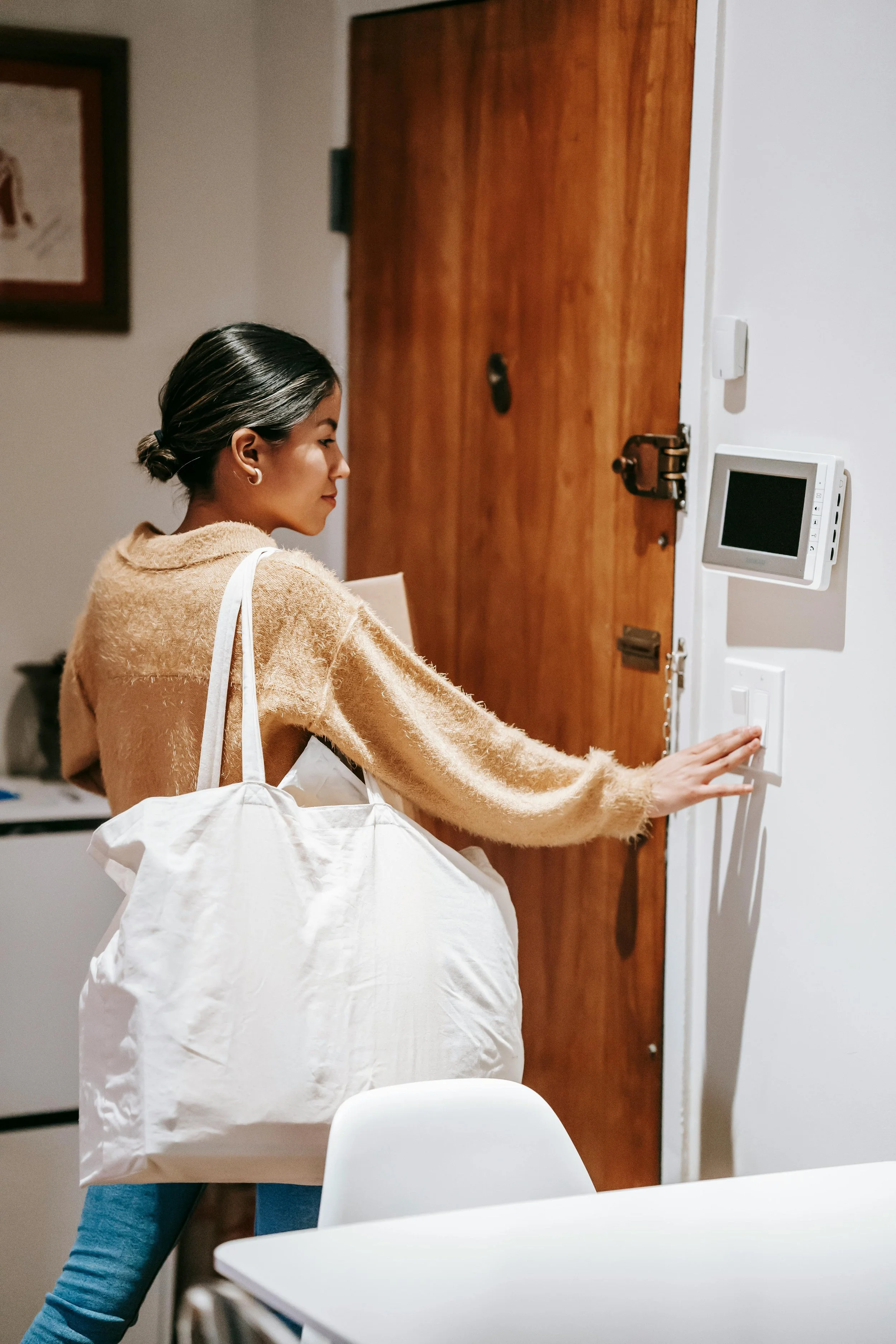 A woman with black hair tied in a low bun wearing a beige fuzzy sweater and blue jeans, standing in an indoor space, reaching towards a white light switch on a wall beside a rounded digital thermostat mounted on the wall.