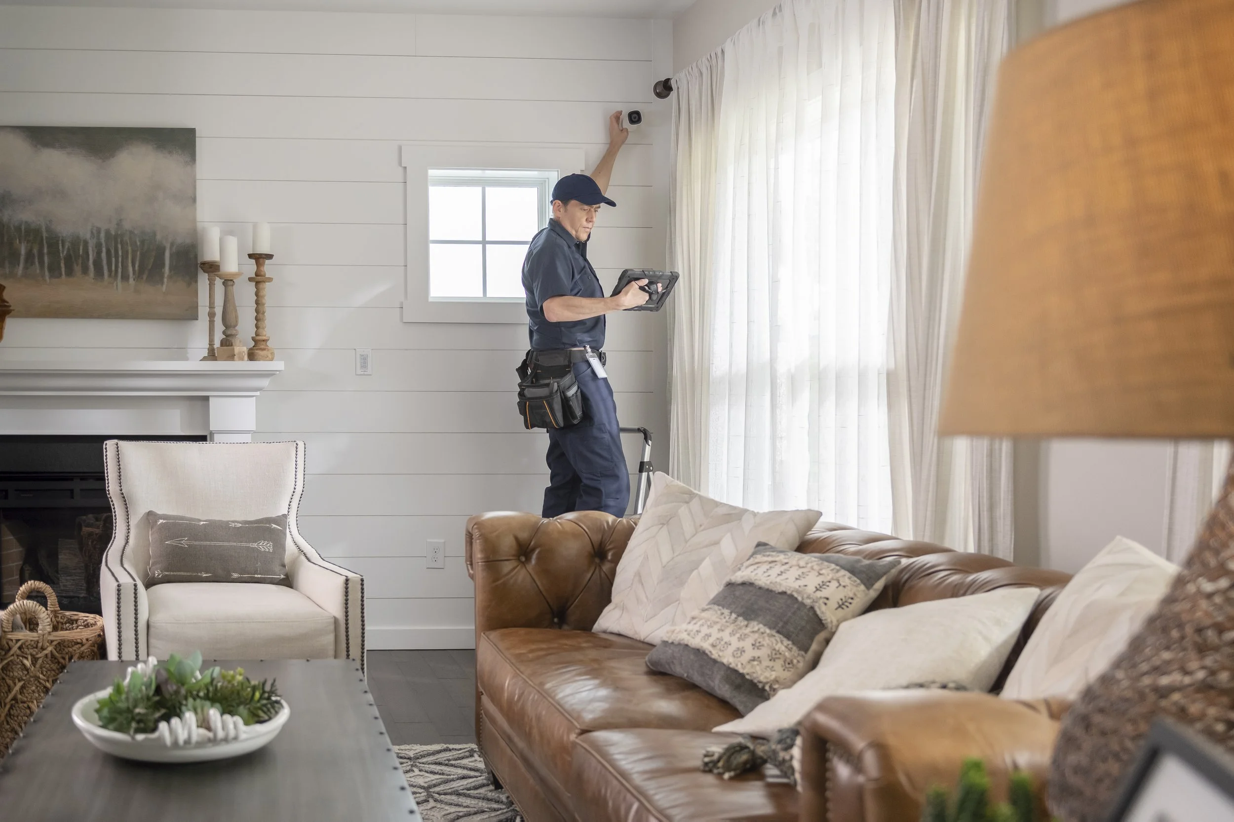A man in a dark blue uniform and cap is installing a security camera near the ceiling of a bright living room, holding a control tablet, with a tool belt around his waist.