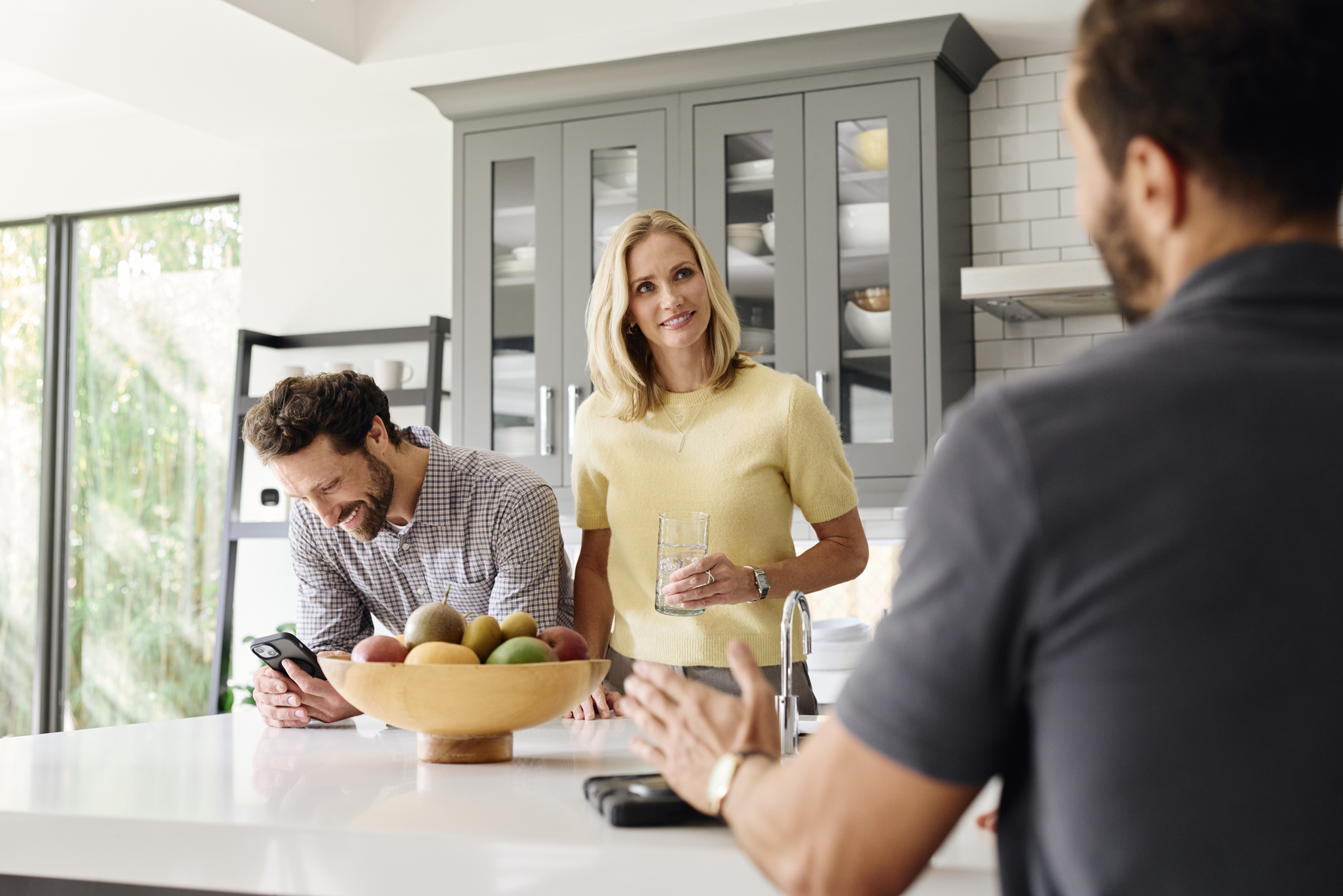 People gathered in a kitchen, engaged in conversation, with a woman holding a glass of water and a bowl of fruit on the counter.