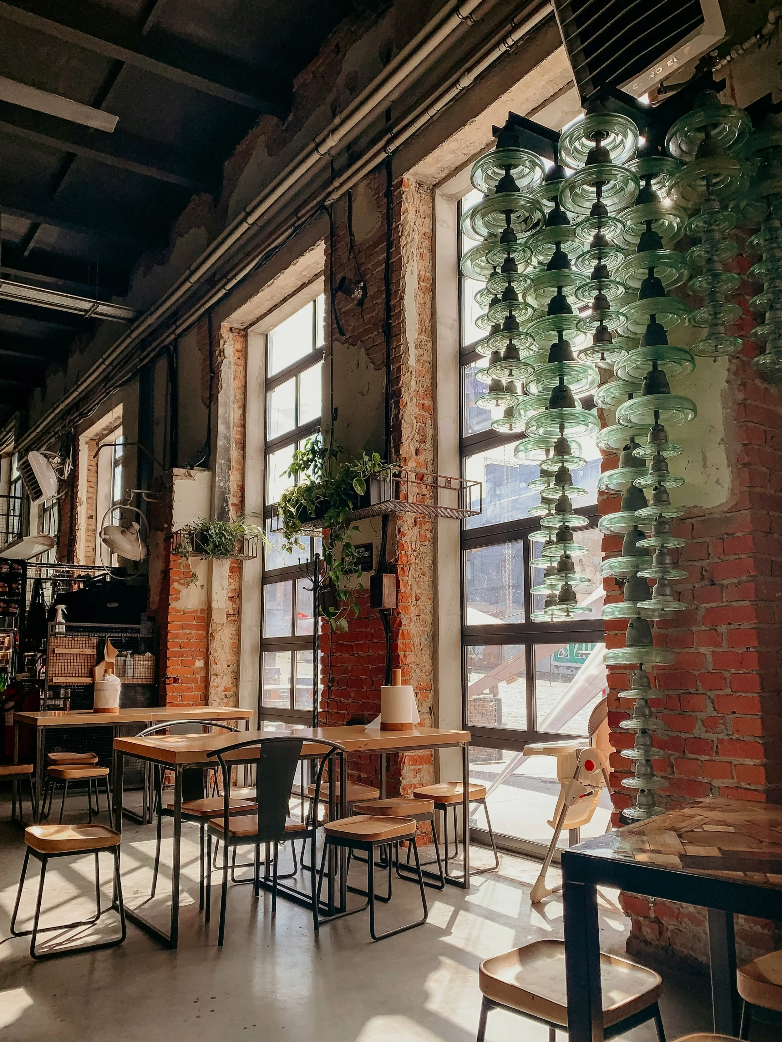 Interior of a café with exposed brick walls, large windows allowing natural light, and modern furniture including wooden tables and chairs. There is a decorative sculpture made from glass plates on the wall near the window, and potted plants hanging near the window.