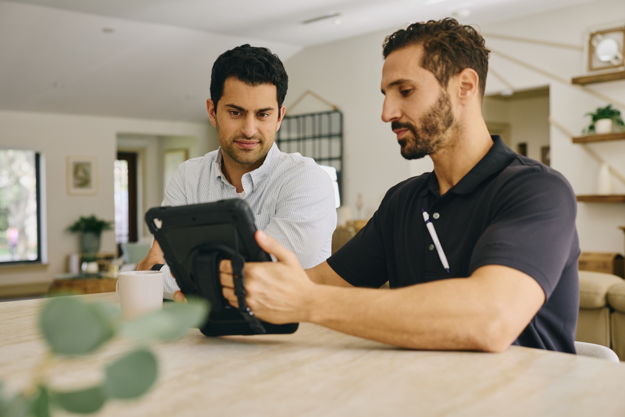 Two men are sitting at a kitchen table looking at a tablet. One man has dark curly hair and is wearing a striped shirt, the other has short curly hair and a beard, wearing a black polo shirt. The background shows a bright kitchen with windows, plants, and wooden shelves.